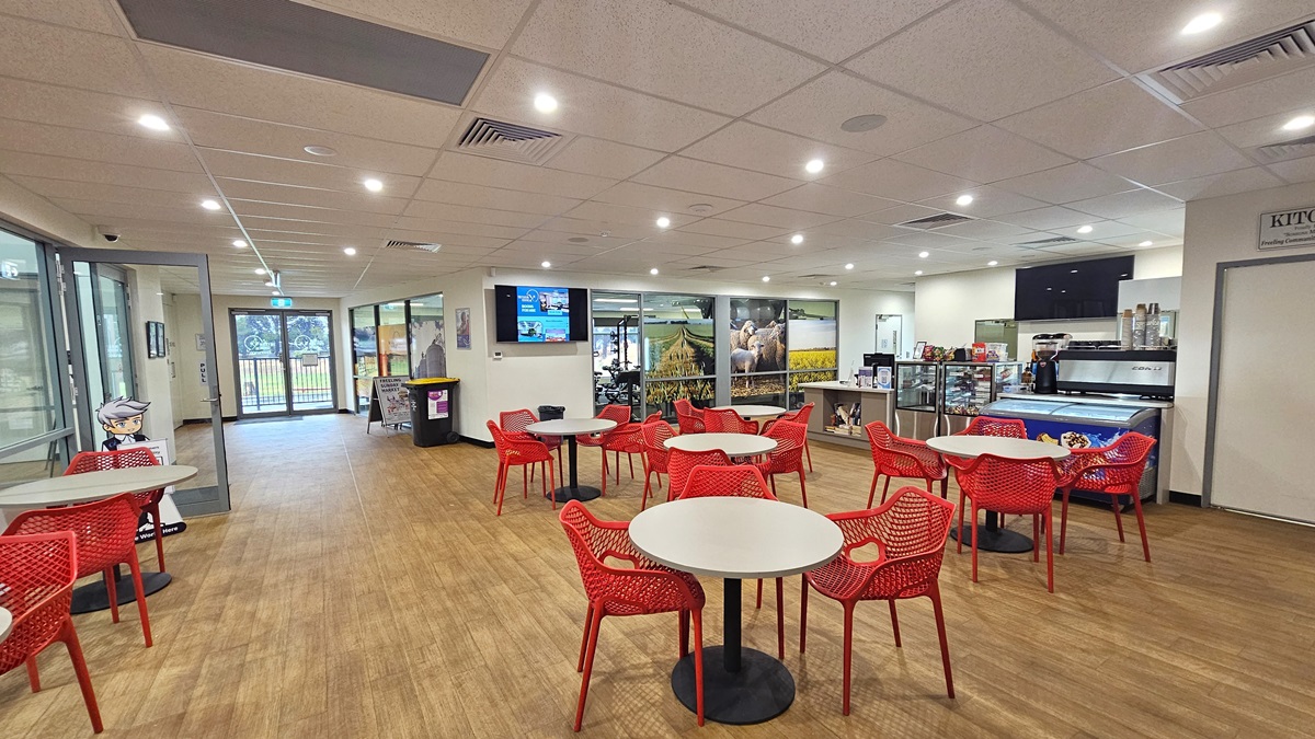 The reception desk, with a cream desk, is on the right. A flat-screen TV is on the wall, and books are on the shelves underneath. Beside it is a display rack showcasing the cafe's items. Movable red chairs and white tables are on the right. Adjacent to the reception is the gym, with a colorful glass wall and a flat-screen. To the left is the entrance to the court. Additional tables and chairs are lined up on the side. The middle wall has a rubbish bin and a free-standing sign. The reception desk, with a cream desk, is on the right. A flat-screen TV is on the wall, and books are on the shelves underneath. Beside it is a display rack showcasing the cafe's items. Movable red chairs and white tables are on the right. Adjacent to the reception is the gym, with a colorful glass wall and a flat-screen. To the left is the entrance to the court. Additional tables and chairs are lined up on the side. The middle wall has a rubbish bin and a free-standing sign.