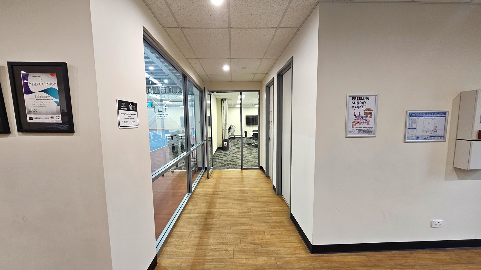 The lobby, with a timber floor, leads to the meeting room equipped with an outward-opening glass door. On the left is a floor-to-ceiling glass wall. On the right are two doors. The other wall has framed posters displaying information about the site's achievements and a venue map. The walls, including the ceiling, are white, with a porous design and circular lights. The lobby, with a timber floor, leads to the meeting room equipped with an outward-opening glass door. On the left is a floor-to-ceiling glass wall. On the right are two doors. The other wall has framed posters displaying information about the site's achievements and a venue map. The walls, including the ceiling, are white, with a porous design and circular lights.