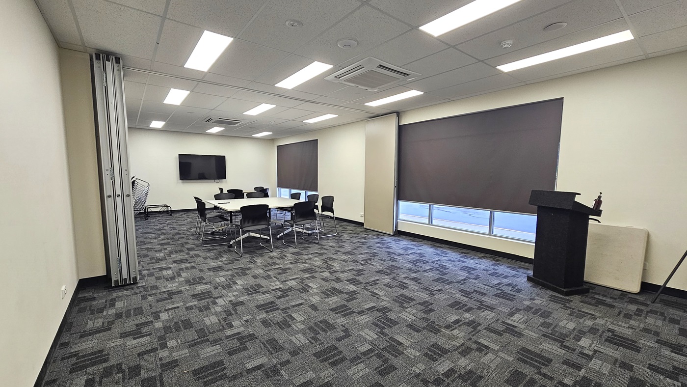 The meeting room features a spacious grey carpet with a checkered design. In the middle, there is a white conference table surrounded by black chairs. At the far end, a flatscreen TV is mounted on the wall. To the right, there are glass windows with black blinds, and a black desk is placed near the wall. On the left, an accordion steel door serves as a separator between two rooms. The ceiling is white with fluorescent lights illuminating the room. The meeting room features a spacious grey carpet with a checkered design. In the middle, there is a white conference table surrounded by black chairs. At the far end, a flatscreen TV is mounted on the wall. To the right, there are glass windows with black blinds, and a black desk is placed near the wall. On the left, an accordion steel door serves as a separator between two rooms. The ceiling is white with fluorescent lights illuminating the room.