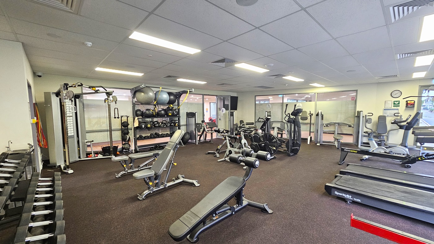 Access to the gym is through a manually operated glass door. A large flatscreen TV is mounted on the left wall near the entrance. The left wall features a floor-to-ceiling glass door adorned with a large poster of a farm landscape. Inside the gym, machines are aligned with rubber matting on the floor and a white ceiling with a porous design and centralized air conditioning. At the far end, large glass windows provide a view of the outside. Access to the gym is through a manually operated glass door. A large flatscreen TV is mounted on the left wall near the entrance. The left wall features a floor-to-ceiling glass door adorned with a large poster of a farm landscape. Inside the gym, machines are aligned with rubber matting on the floor and a white ceiling with a porous design and centralized air conditioning. At the far end, large glass windows provide a view of the outside.