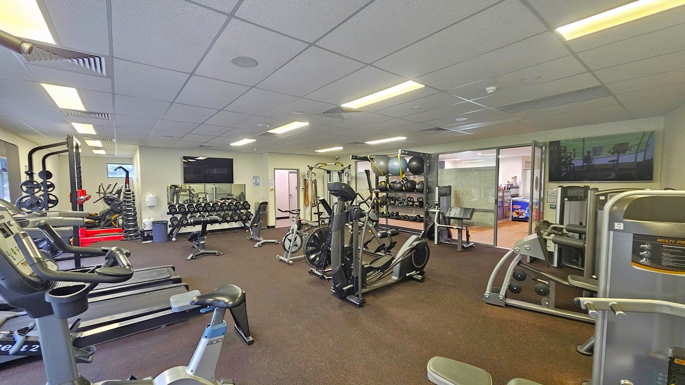 On the left, there are treadmills, stationary bikes, and Smith machines. In the middle, weights are neatly arranged in a display rack, and a flatscreen TV is mounted on the white wall. On the right, a display rack holds stability balls, medicine balls, weights, and kettlebells. Other machines are placed on the right near the door, along with a flat-screen TV. The ceiling is white with air conditioning vents. On the left, there are treadmills, stationary bikes, and Smith machines. In the middle, weights are neatly arranged in a display rack, and a flatscreen TV is mounted on the white wall. On the right, a display rack holds stability balls, medicine balls, weights, and kettlebells. Other machines are placed on the right near the door, along with a flat-screen TV. The ceiling is white with air conditioning vents.