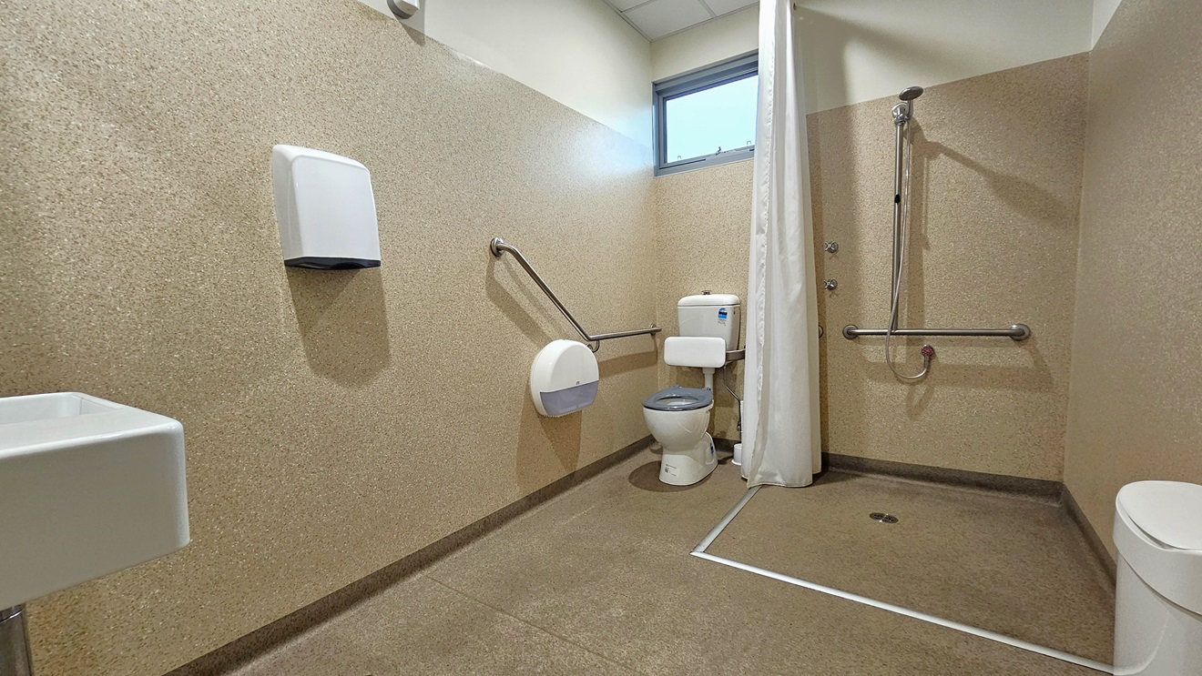 Inside the bathroom, brown granite walls enclose the space. On the left, a wash basin and hand dryer are mounted on the wall. In the corner, the toilet bowl is equipped with hand railings. A glass window is placed overhead. In the middle, a shower is mounted to the wall, separated by a white shower curtain. On the right is a rubbish bin. Inside the bathroom, brown granite walls enclose the space. On the left, a wash basin and hand dryer are mounted on the wall. In the corner, the toilet bowl is equipped with hand railings. A glass window is placed overhead. In the middle, a shower is mounted to the wall, separated by a white shower curtain. On the right is a rubbish bin.