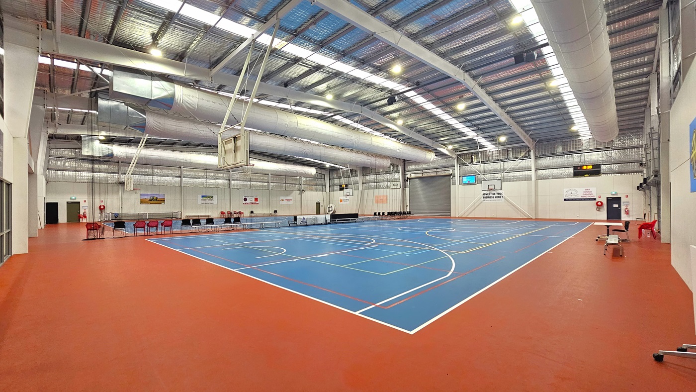 Access to the stadium is through a manual, outward-opening glass door equipped with a non-slip mat. On the right is a framed poster displaying the site's achievements. To the left is a table with a red plastic chair. Inside the stadium, a large and open space, the size of two basketball courts marked for various ball games. The ceiling is adorned with exposed white beams, reflective silver insulation, and a mixture of natural skylights and spotlights. Access to the stadium is through a manual, outward-opening glass door equipped with a non-slip mat. On the right is a framed poster displaying the site's achievements. To the left is a table with a red plastic chair. Inside the stadium, a large and open space, the size of two basketball courts marked for various ball games. The ceiling is adorned with exposed white beams, reflective silver insulation, and a mixture of natural skylights and spotlights.