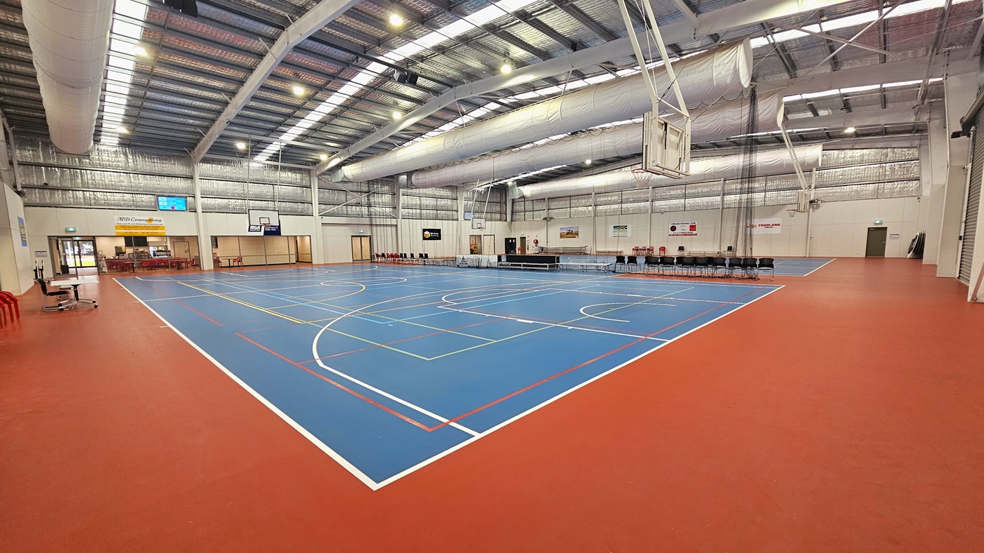The solid floor exhibits a combination of blue and red, with markings for various ball games. The ceiling showcases exposed white beams, air conditioning vents, reflective silver insulation, and a blend of natural skylights and spotlights. In the middle, chairs and tables are arranged to distinguish the two courts. Digital screens are affixed to the left walls, and tables and chairs are positioned on the left. Basketball boards are installed on both sides. The solid floor exhibits a combination of blue and red, with markings for various ball games. The ceiling showcases exposed white beams, air conditioning vents, reflective silver insulation, and a blend of natural skylights and spotlights. In the middle, chairs and tables are arranged to distinguish the two courts. Digital screens are affixed to the left walls, and tables and chairs are positioned on the left. Basketball boards are installed on both sides.