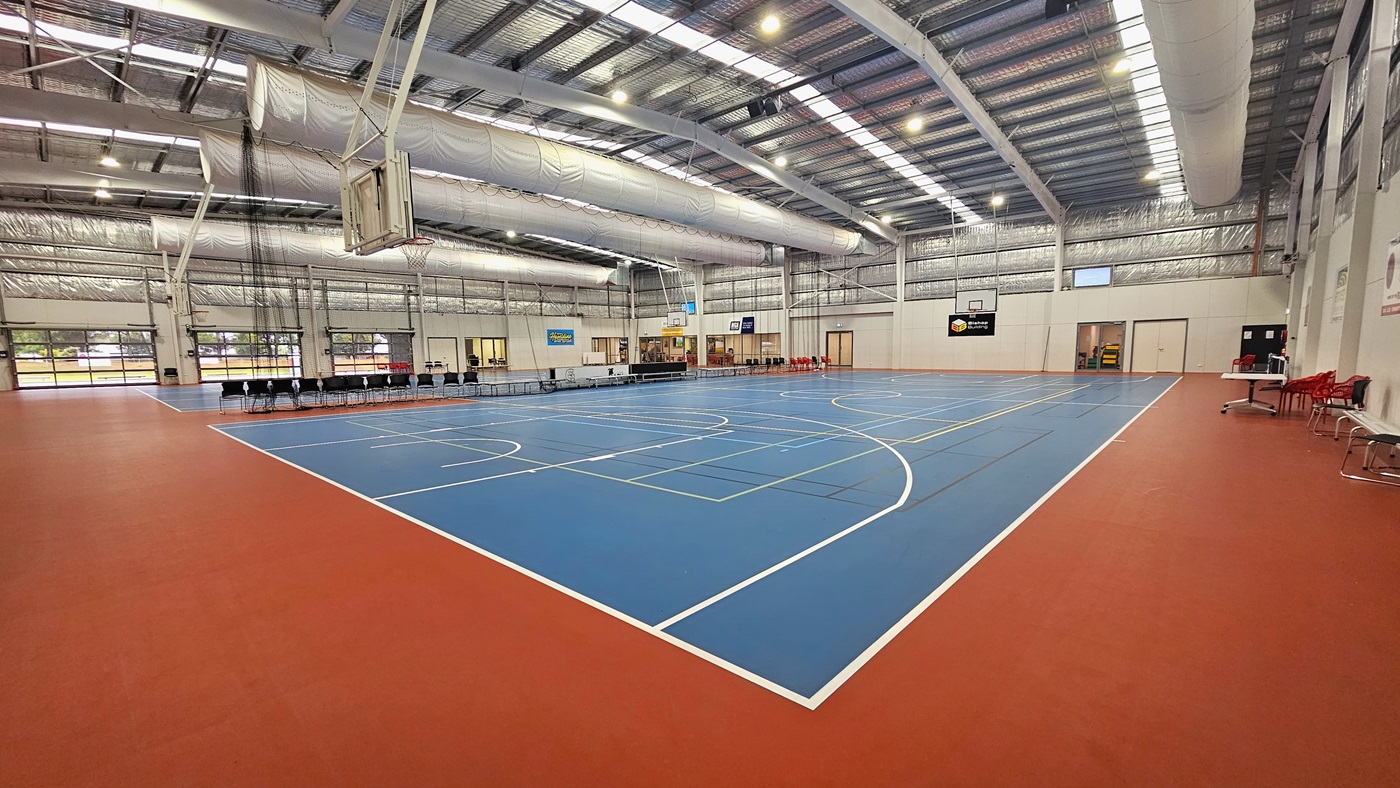 The floor features a blend of blue and red with markings for various ball games. The ceiling displays exposed white beams, air conditioning vents, reflective silver insulation, and a mix of natural skylights and spotlights. In the middle, chairs and tables are arranged to separate the two courts. Digital screens are mounted on the middle walls. Basketball boards are positioned on both sides. On the left, tables and chairs are placed near the white wall. The floor features a blend of blue and red with markings for various ball games. The ceiling displays exposed white beams, air conditioning vents, reflective silver insulation, and a mix of natural skylights and spotlights. In the middle, chairs and tables are arranged to separate the two courts. Digital screens are mounted on the middle walls. Basketball boards are positioned on both sides. On the left, tables and chairs are placed near the white wall.