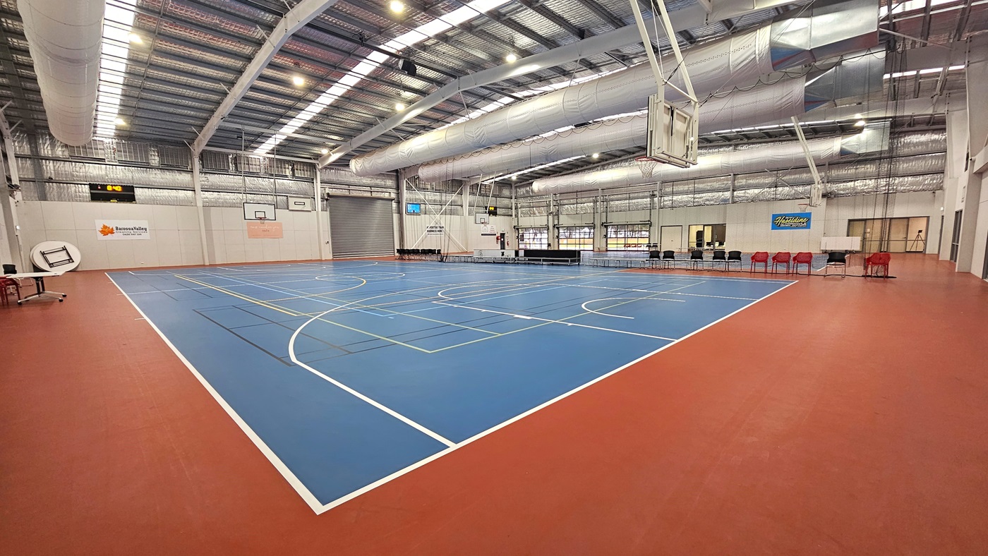 The solid floor exhibits a combination of blue and red, with markings for various ball games. The ceiling showcases exposed white beams, air conditioning vents, reflective silver insulation, and a blend of natural skylights and spotlights. In the middle, chairs and tables are arranged to distinguish the two courts. Digital screens are affixed to the middle walls. Basketball boards are installed on both sides.In the left, tables and chairs are positioned near the white wall. The solid floor exhibits a combination of blue and red, with markings for various ball games. The ceiling showcases exposed white beams, air conditioning vents, reflective silver insulation, and a blend of natural skylights and spotlights. In the middle, chairs and tables are arranged to distinguish the two courts. Digital screens are affixed to the middle walls. Basketball boards are installed on both sides.In the left, tables and chairs are positioned near the white wall.