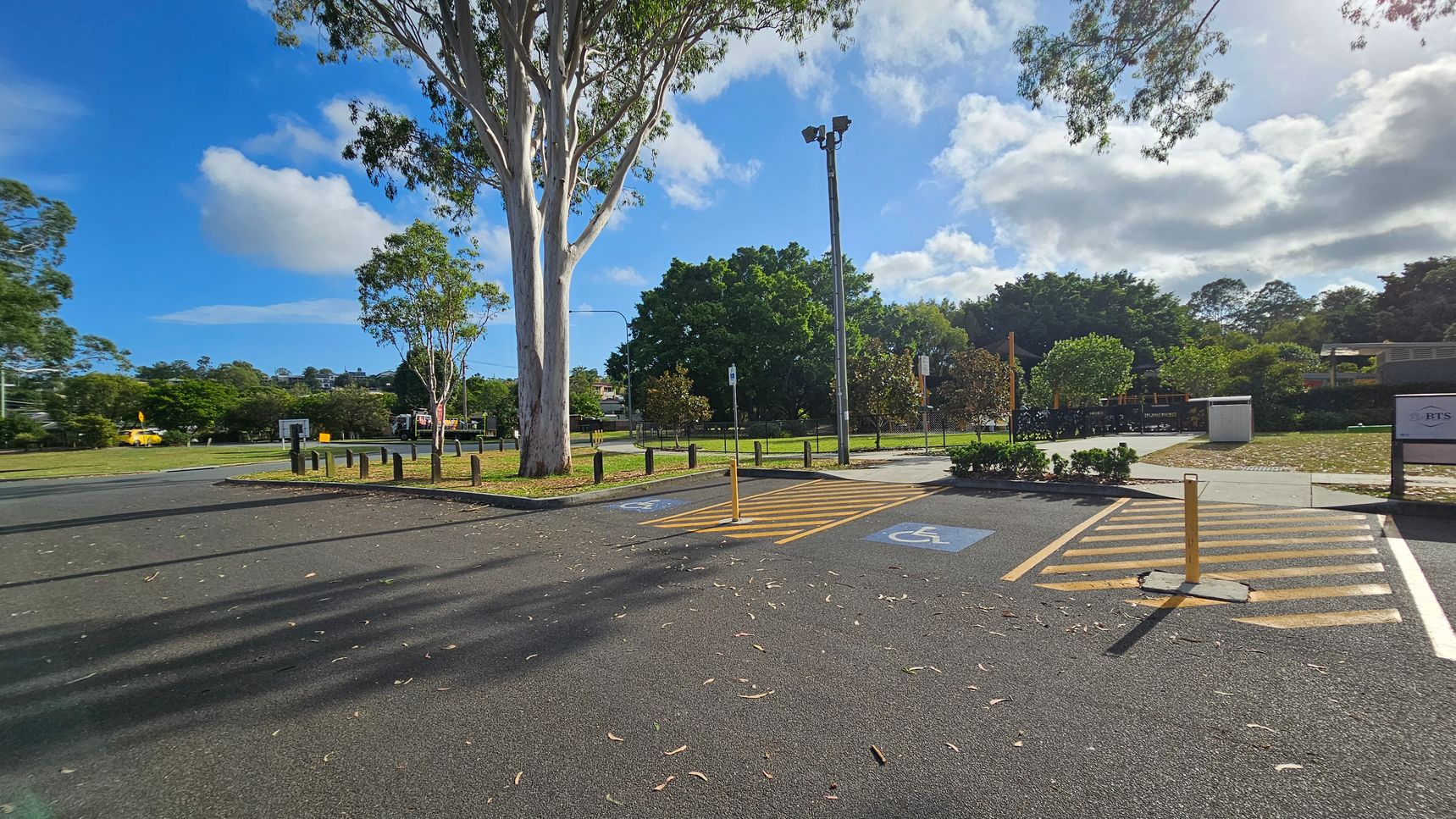 A car park with two marked disabled parking spaces in the foreground, featuring blue wheelchair symbols. A large tree stands to the left, casting shadows on the asphalt. In the background, there are more trees and a grassy area, with a fence and a small structure visible. A tall light pole is near the centre, and a sign is on the right. The ground is scattered with leaves and debris. A car park with two marked disabled parking spaces in the foreground, featuring blue wheelchair symbols. A large tree stands to the left, casting shadows on the asphalt. In the background, there are more trees and a grassy area, with a fence and a small structure visible. A tall light pole is near the centre, and a sign is on the right. The ground is scattered with leaves and debris.