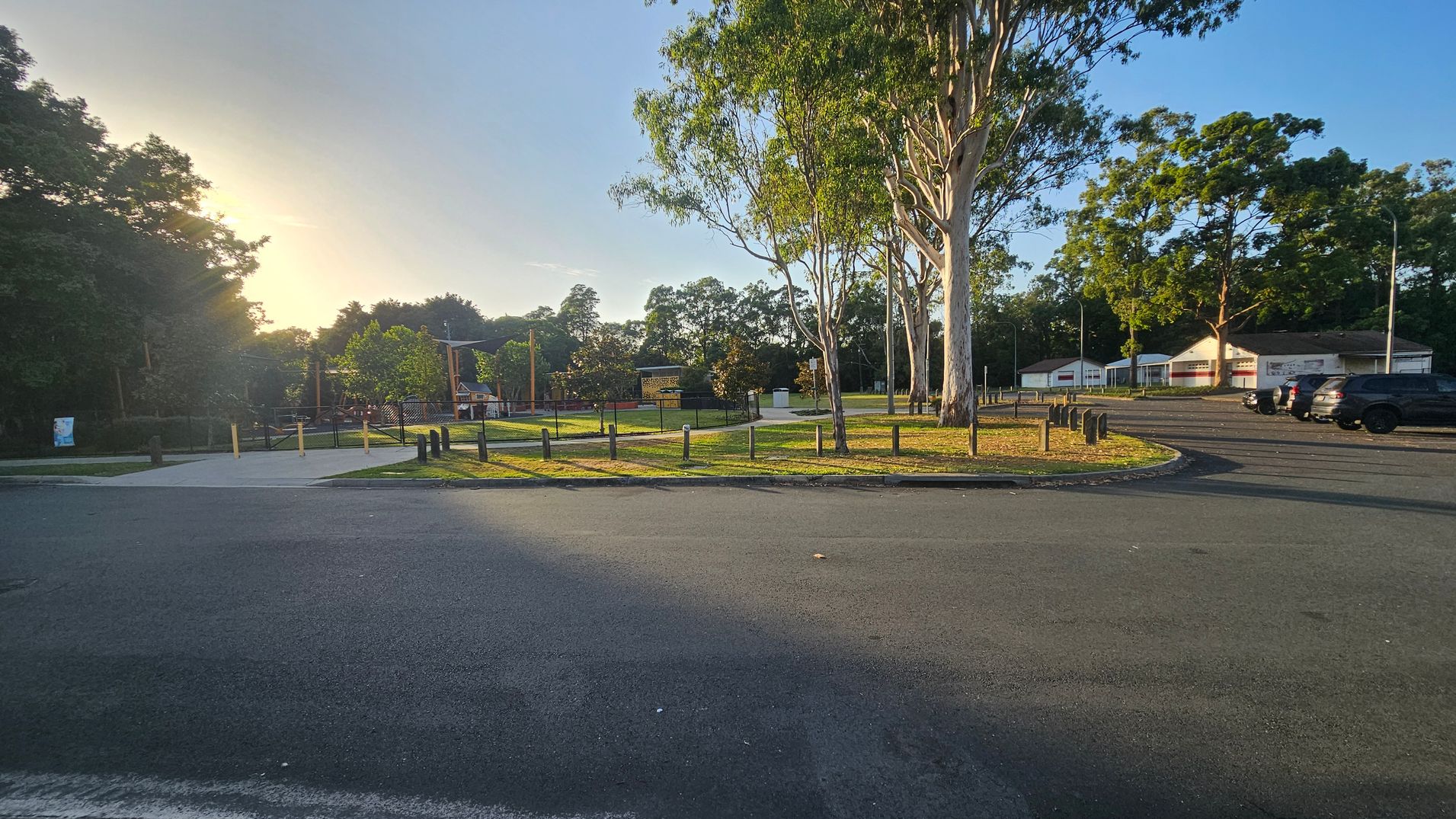 A park with a paved path and grassy area in the centre, surrounded by tall trees. On the right, there are several parked cars and a few buildings with white walls and red signage. The left side has more trees and a fenced area. The ground is a mix of asphalt and grass, and the sun is setting in the background, casting long shadows. A park with a paved path and grassy area in the centre, surrounded by tall trees. On the right, there are several parked cars and a few buildings with white walls and red signage. The left side has more trees and a fenced area. The ground is a mix of asphalt and grass, and the sun is setting in the background, casting long shadows.