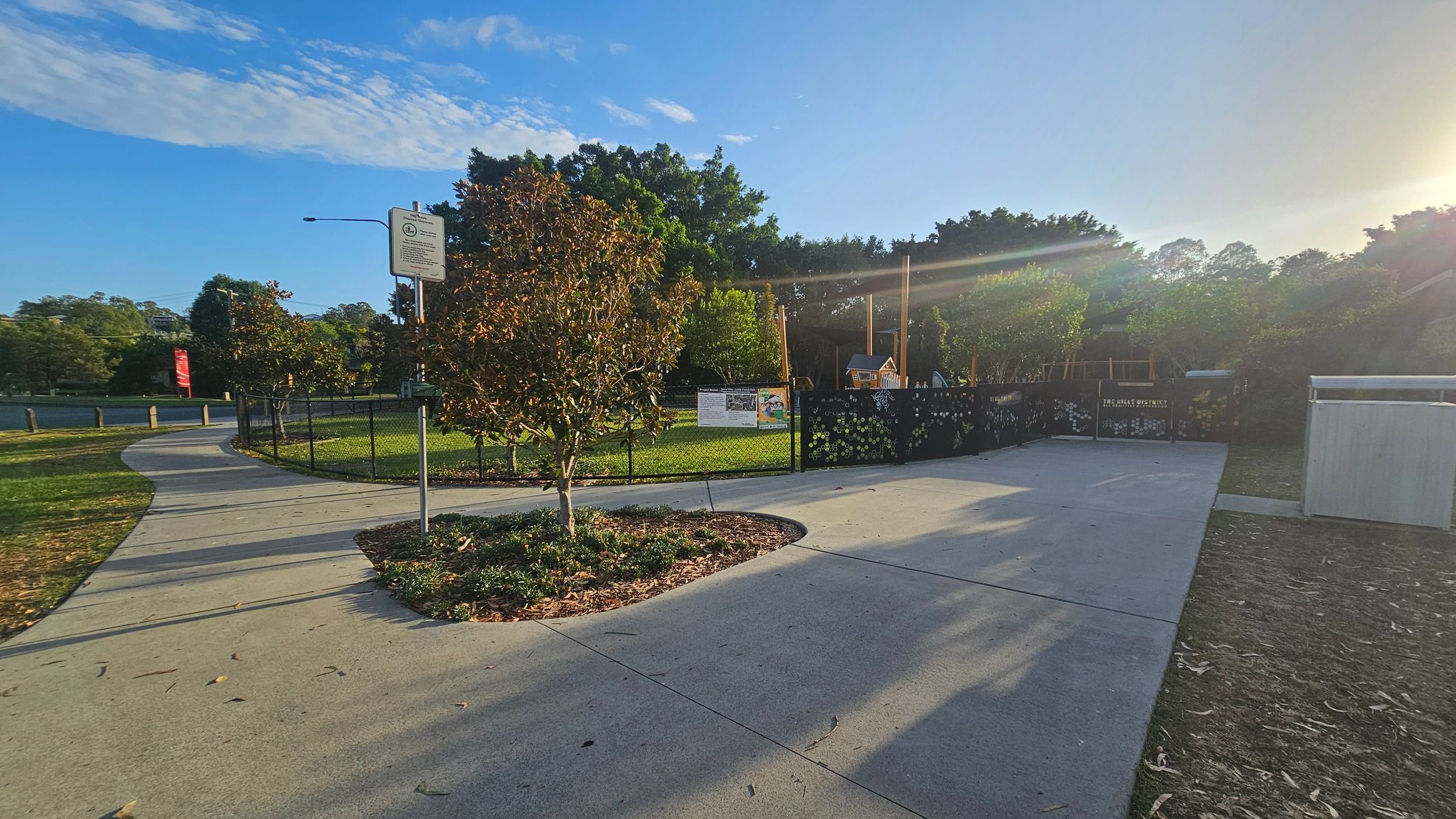 A paved pathway curves to the left, bordered by grass and small trees. A signpost stands near the path. To the right, a fenced area encloses a playground with a small playhouse and equipment. The fence has decorative elements and a sign. Trees and greenery fill the background, with sunlight streaming from the right, casting long shadows on the ground. The sky is clear with some clouds. A paved pathway curves to the left, bordered by grass and small trees. A signpost stands near the path. To the right, a fenced area encloses a playground with a small playhouse and equipment. The fence has decorative elements and a sign. Trees and greenery fill the background, with sunlight streaming from the right, casting long shadows on the ground. The sky is clear with some clouds.