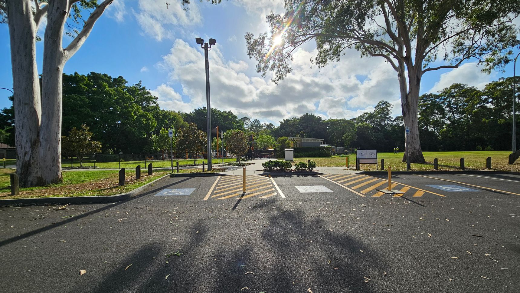 A car park with designated disabled parking spaces marked in blue and yellow lines in the foreground. Two large trees stand on either side, casting shadows on the asphalt. In the background, a grassy area with more trees and a playground is visible. A tall light pole is centrally located, and the sun shines through the clouds, creating a bright spot in the sky. A sign is visible near the trees on the right. A car park with designated disabled parking spaces marked in blue and yellow lines in the foreground. Two large trees stand on either side, casting shadows on the asphalt. In the background, a grassy area with more trees and a playground is visible. A tall light pole is centrally located, and the sun shines through the clouds, creating a bright spot in the sky. A sign is visible near the trees on the right.