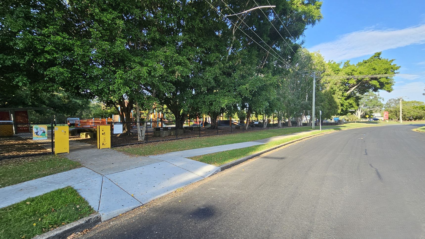 A quiet street with a wide road on the right and a footpath on the left. The left side features a park enclosed by a black fence, with large green trees providing shade. Yellow entry gates and signs are visible at the park entrance. The park contains playground equipment in the background. Power lines run overhead, and a grassy verge separates the footpath from the road. A quiet street with a wide road on the right and a footpath on the left. The left side features a park enclosed by a black fence, with large green trees providing shade. Yellow entry gates and signs are visible at the park entrance. The park contains playground equipment in the background. Power lines run overhead, and a grassy verge separates the footpath from the road.
