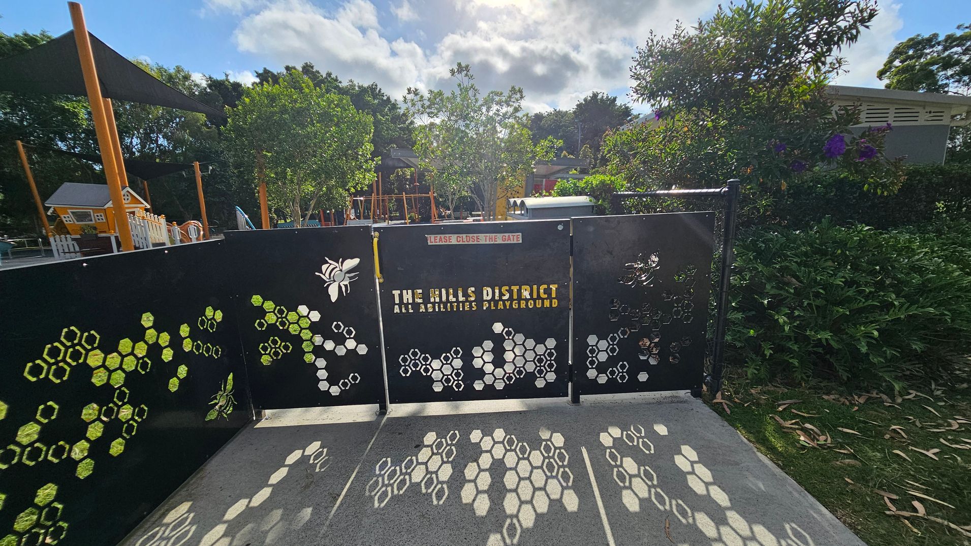 A black gate with honeycomb and bee designs, labelled "The Hills District All Abilities Playground," stands in the foreground. The ground is concrete with honeycomb patterns. Behind the gate, there are trees and playground equipment, including a small house structure and slides. The background is filled with lush greenery and a partly cloudy sky. A black gate with honeycomb and bee designs, labelled "The Hills District All Abilities Playground," stands in the foreground. The ground is concrete with honeycomb patterns. Behind the gate, there are trees and playground equipment, including a small house structure and slides. The background is filled with lush greenery and a partly cloudy sky.