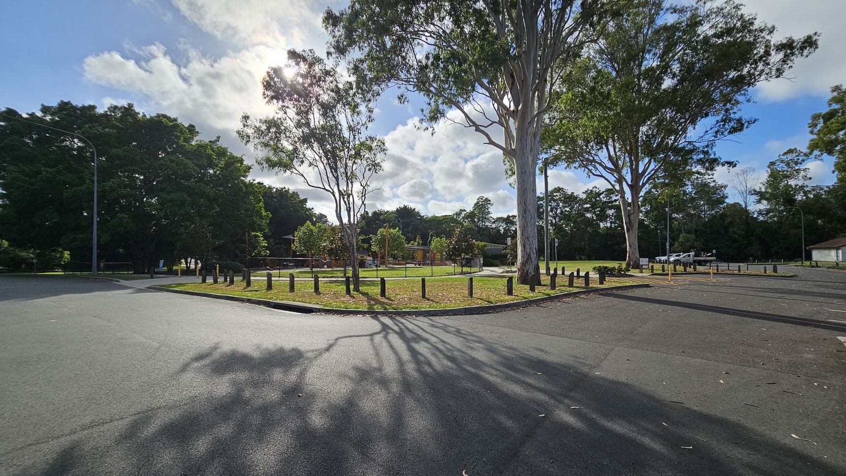 A park with a grassy area in the centre, surrounded by a low wooden barrier. Tall trees with sparse leaves are scattered throughout, casting shadows on the ground. To the right, a paved parking area with a few cars is visible. On the left, dense foliage forms a backdrop. The sky is partly cloudy, and the sun is partially obscured by the trees. The road in the foreground is smooth and dark, with tree shadows stretching across it. A park with a grassy area in the centre, surrounded by a low wooden barrier. Tall trees with sparse leaves are scattered throughout, casting shadows on the ground. To the right, a paved parking area with a few cars is visible. On the left, dense foliage forms a backdrop. The sky is partly cloudy, and the sun is partially obscured by the trees. The road in the foreground is smooth and dark, with tree shadows stretching across it.