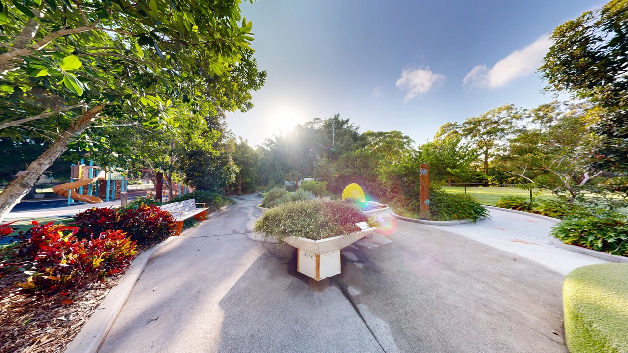 A garden scene with large, hexagonal planters filled with various plants and flowers on the left. In the centre, a pathway leads through the garden, with colourful, dome-shaped structures resembling beehives on the right. The background is filled with lush green trees and a glimpse of a playground with a slide. The ground is paved, and the area is well-maintained, with vibrant greenery throughout. A garden scene with large, hexagonal planters filled with various plants and flowers on the left. In the centre, a pathway leads through the garden, with colourful, dome-shaped structures resembling beehives on the right. The background is filled with lush green trees and a glimpse of a playground with a slide. The ground is paved, and the area is well-maintained, with vibrant greenery throughout.