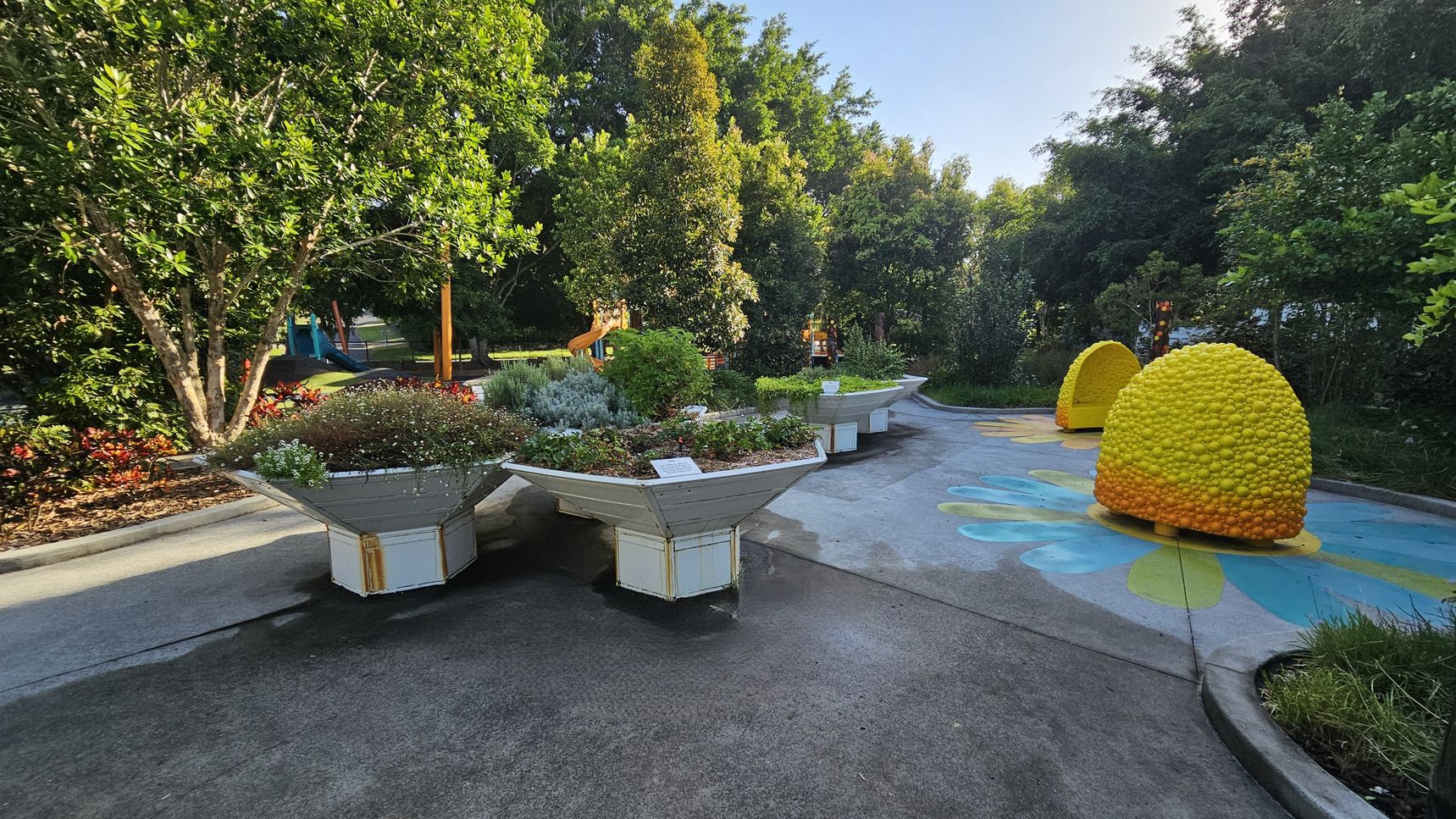 A garden scene with large, hexagonal planters filled with various plants and flowers on the left. In the centre, a pathway leads through the garden, with colourful, dome-shaped structures resembling beehives on the right. The background is filled with lush green trees and a glimpse of a playground with a slide. The ground is paved, and the area is well-maintained, with vibrant greenery throughout. A garden scene with large, hexagonal planters filled with various plants and flowers on the left. In the centre, a pathway leads through the garden, with colourful, dome-shaped structures resembling beehives on the right. The background is filled with lush green trees and a glimpse of a playground with a slide. The ground is paved, and the area is well-maintained, with vibrant greenery throughout.