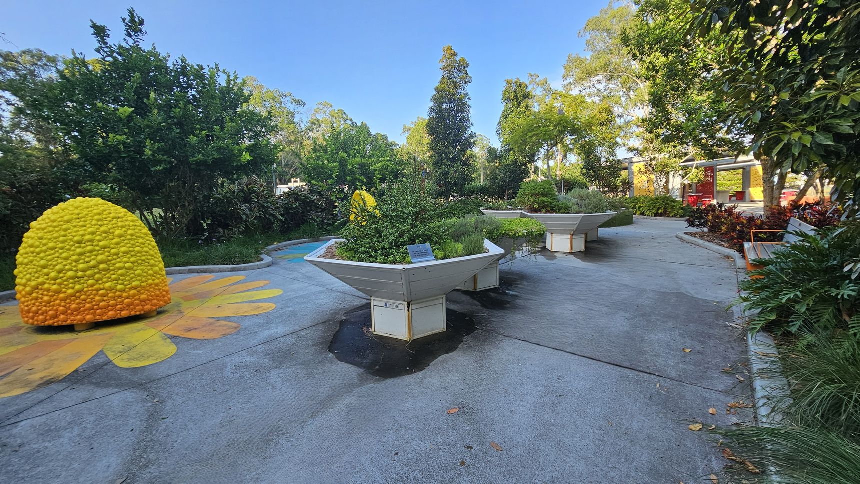 A garden area with a concrete path. On the left, there's a large, dome-shaped sculpture resembling a beehive, painted in yellow and orange. In the centre, there are large, hexagonal planters filled with various green plants. To the right, a wooden bench is surrounded by lush foliage. In the background, trees and shrubs create a dense, green backdrop, and a structure with red accents is visible. The ground is decorated with colourful patterns. A garden area with a concrete path. On the left, there's a large, dome-shaped sculpture resembling a beehive, painted in yellow and orange. In the centre, there are large, hexagonal planters filled with various green plants. To the right, a wooden bench is surrounded by lush foliage. In the background, trees and shrubs create a dense, green backdrop, and a structure with red accents is visible. The ground is decorated with colourful patterns.