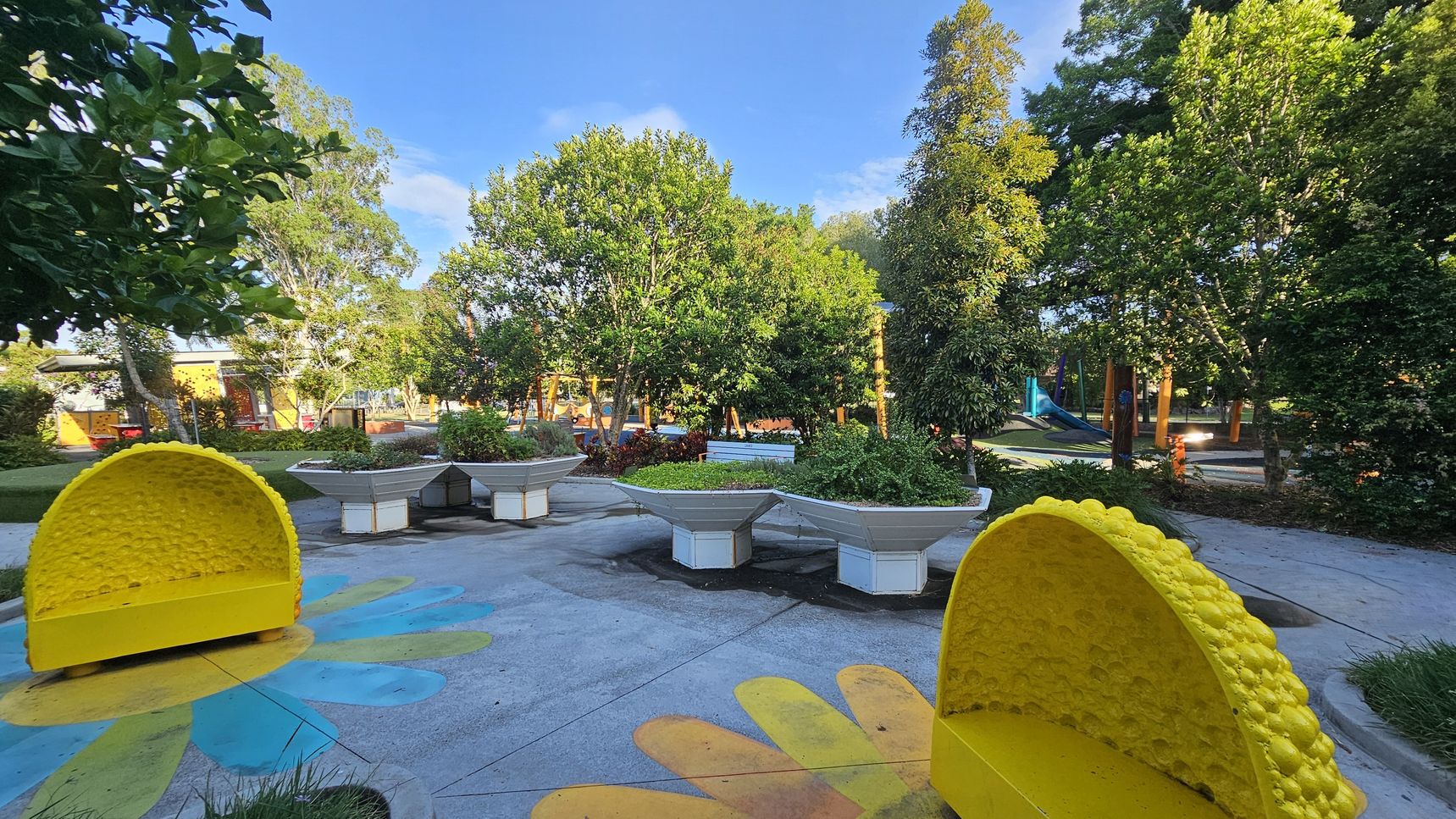 A vibrant park scene with two large, yellow, semi-circular benches resembling lemon slices on the left and right. The ground features colourful flower patterns. In the centre, there are several large, white, hexagonal planters filled with greenery. The background is filled with lush trees and a playground area with slides and climbing structures. The sky is clear and blue, and the area is well-lit, suggesting a sunny day. A vibrant park scene with two large, yellow, semi-circular benches resembling lemon slices on the left and right. The ground features colourful flower patterns. In the centre, there are several large, white, hexagonal planters filled with greenery. The background is filled with lush trees and a playground area with slides and climbing structures. The sky is clear and blue, and the area is well-lit, suggesting a sunny day.