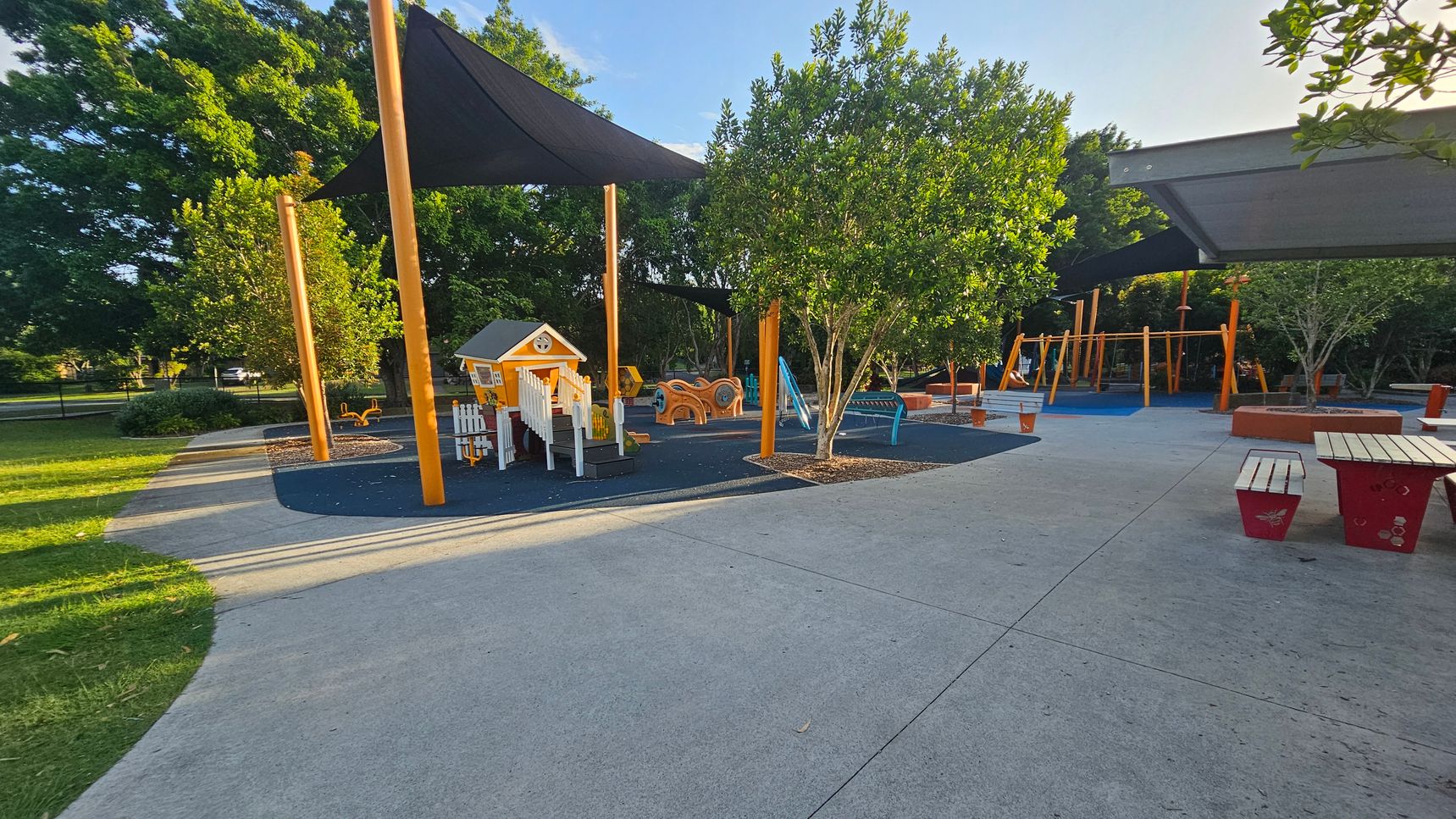 A playground with a small playhouse and various play equipment on the left, surrounded by trees and shaded by large triangular canopies. The ground is covered with a mix of soft play surfaces and concrete paths. On the right, there are picnic tables with red benches. The background features more trees and additional play structures, including swings. The area is well-maintained and spacious, with a mix of sun and shade. A playground with a small playhouse and various play equipment on the left, surrounded by trees and shaded by large triangular canopies. The ground is covered with a mix of soft play surfaces and concrete paths. On the right, there are picnic tables with red benches. The background features more trees and additional play structures, including swings. The area is well-maintained and spacious, with a mix of sun and shade.