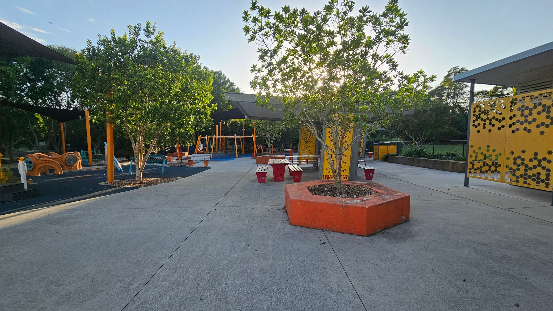 A playground with a concrete path in the foreground. On the left, there are trees and playground equipment with orange and blue structures. In the centre, a tree is planted in an orange hexagonal planter, surrounded by benches and red stools. On the right, a yellow panel with a honeycomb pattern is visible, along with more trees and a grassy area in the background. The area is shaded by large canopies. A playground with a concrete path in the foreground. On the left, there are trees and playground equipment with orange and blue structures. In the centre, a tree is planted in an orange hexagonal planter, surrounded by benches and red stools. On the right, a yellow panel with a honeycomb pattern is visible, along with more trees and a grassy area in the background. The area is shaded by large canopies.