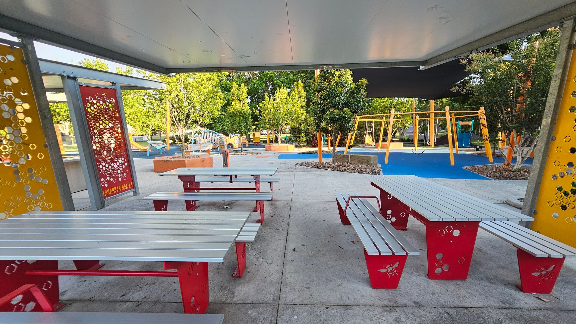 Under a grey ceiling, there are several picnic tables with red metal legs and grey tops. The floor is concrete. In the background, a playground features swings and climbing structures on a blue rubber surface. Trees and shrubs are scattered throughout the area. Decorative panels with hexagonal patterns and bee motifs are on the left and right, with one labelled "Sugarbag Native Bee." Under a grey ceiling, there are several picnic tables with red metal legs and grey tops. The floor is concrete. In the background, a playground features swings and climbing structures on a blue rubber surface. Trees and shrubs are scattered throughout the area. Decorative panels with hexagonal patterns and bee motifs are on the left and right, with one labelled "Sugarbag Native Bee."