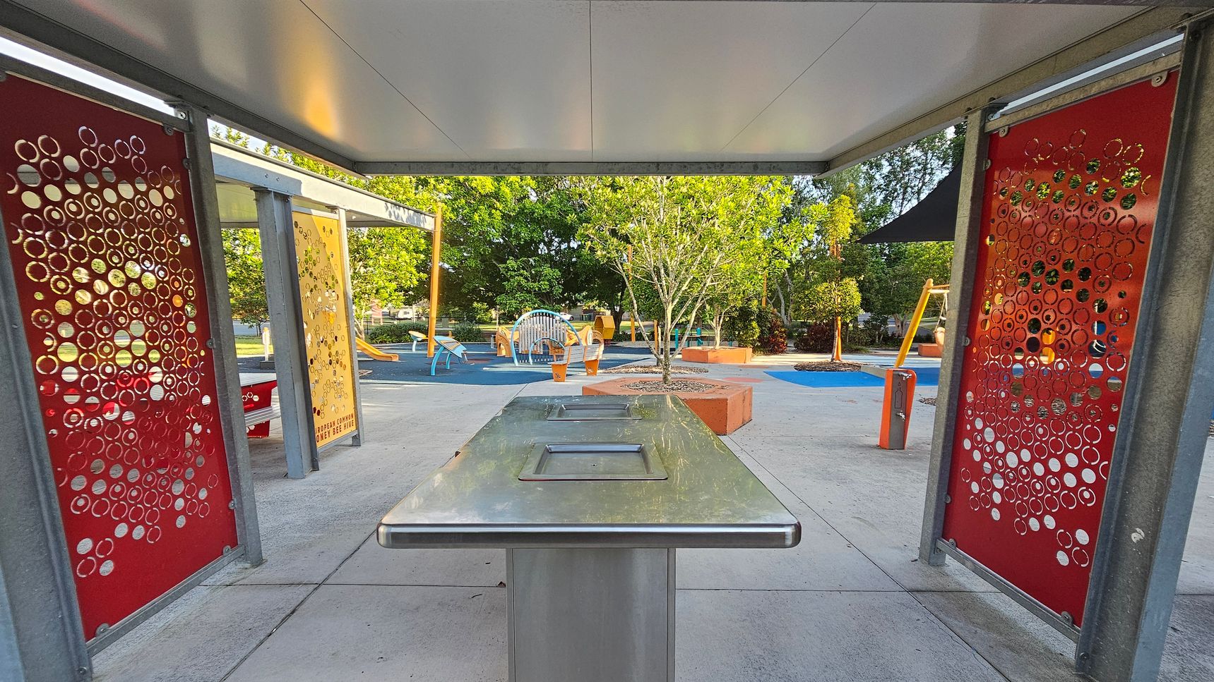 A covered outdoor area with a stainless steel table in the centre. On the left and right are red panels with circular cut-out patterns. In the background, a playground with various equipment, including a slide and climbing structures, is visible. Trees and shrubs are scattered throughout, with some planted in orange-bordered planters. The ground is a mix of concrete and blue rubberised surfaces. The ceiling is flat and grey. A covered outdoor area with a stainless steel table in the centre. On the left and right are red panels with circular cut-out patterns. In the background, a playground with various equipment, including a slide and climbing structures, is visible. Trees and shrubs are scattered throughout, with some planted in orange-bordered planters. The ground is a mix of concrete and blue rubberised surfaces. The ceiling is flat and grey.