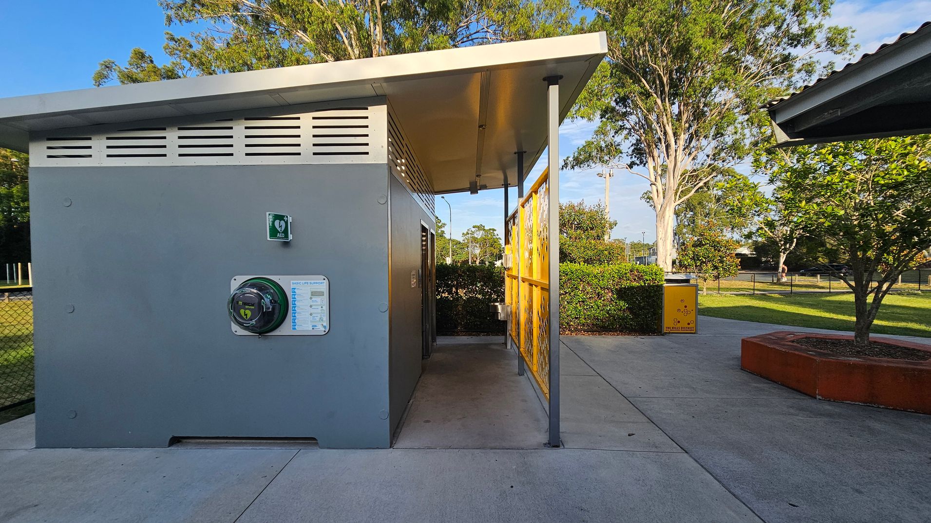 A small grey building with a slanted roof is on the left, featuring an automated external defibrillator (AED) mounted on the wall. A yellow decorative panel is on the right side of the building. In the background, there are trees and a grassy area with a yellow bin. The ground is paved with concrete, and a tree is planted in a red circular planter on the right. A small grey building with a slanted roof is on the left, featuring an automated external defibrillator (AED) mounted on the wall. A yellow decorative panel is on the right side of the building. In the background, there are trees and a grassy area with a yellow bin. The ground is paved with concrete, and a tree is planted in a red circular planter on the right.