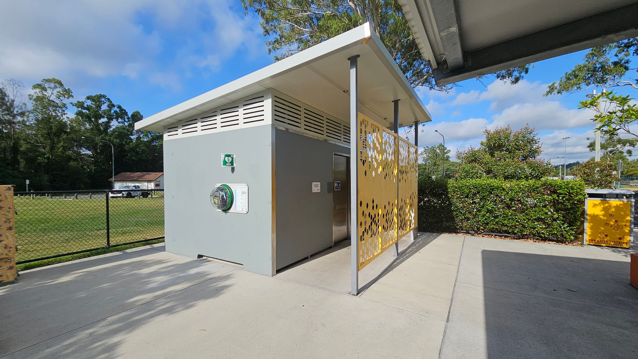 A small, modern public toilet structure with a grey exterior and a slanted white roof. On the right, there's a decorative yellow panel with circular cutouts. A defibrillator is mounted on the wall. In the background, a grassy field is enclosed by a black chain-link fence, with a parked white vehicle and a small building visible. Bushes and trees are scattered around, and a yellow bin is on the right. The ground is paved with concrete. A small, modern public toilet structure with a grey exterior and a slanted white roof. On the right, there's a decorative yellow panel with circular cutouts. A defibrillator is mounted on the wall. In the background, a grassy field is enclosed by a black chain-link fence, with a parked white vehicle and a small building visible. Bushes and trees are scattered around, and a yellow bin is on the right. The ground is paved with concrete.