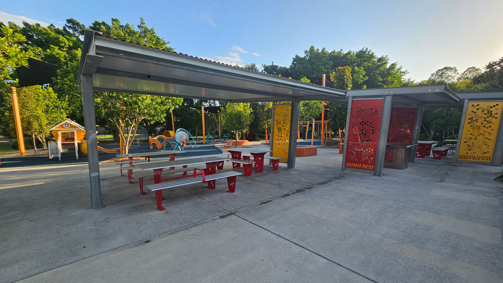 A park area with a covered seating section featuring metal picnic tables with red legs and benches. The shelter has a corrugated metal roof. To the right, there are decorative panels in red and yellow with circular cutouts. In the background, a playground with various equipment, including a small playhouse and climbing structures, is visible. The area is surrounded by trees, and the ground is paved with concrete. A park area with a covered seating section featuring metal picnic tables with red legs and benches. The shelter has a corrugated metal roof. To the right, there are decorative panels in red and yellow with circular cutouts. In the background, a playground with various equipment, including a small playhouse and climbing structures, is visible. The area is surrounded by trees, and the ground is paved with concrete.