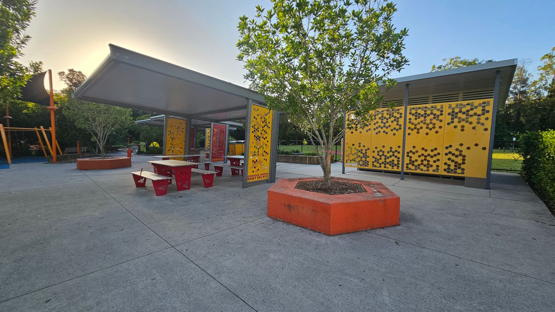 A playground area with a concrete floor. On the left, there's a play structure with orange supports. In the centre, a covered seating area with red benches and tables is visible. A tree is planted in an orange hexagonal planter. On the right, a structure with yellow panels featuring a honeycomb pattern. In the background, there are trees and a grassy area enclosed by a fence. A playground area with a concrete floor. On the left, there's a play structure with orange supports. In the centre, a covered seating area with red benches and tables is visible. A tree is planted in an orange hexagonal planter. On the right, a structure with yellow panels featuring a honeycomb pattern. In the background, there are trees and a grassy area enclosed by a fence.