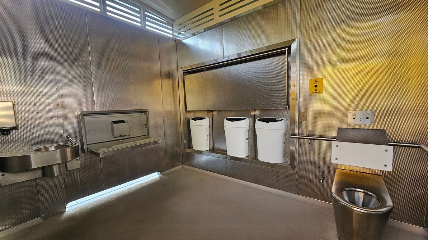 A public restroom with metallic walls and a concrete floor. On the left, a stainless steel sink with a soap dispenser is mounted on the wall. In the centre, a baby changing station is above three white bins. On the right, a stainless steel toilet is visible, with a sharps disposal box mounted above it. The ceiling has a vented design, allowing light to filter through. A public restroom with metallic walls and a concrete floor. On the left, a stainless steel sink with a soap dispenser is mounted on the wall. In the centre, a baby changing station is above three white bins. On the right, a stainless steel toilet is visible, with a sharps disposal box mounted above it. The ceiling has a vented design, allowing light to filter through.