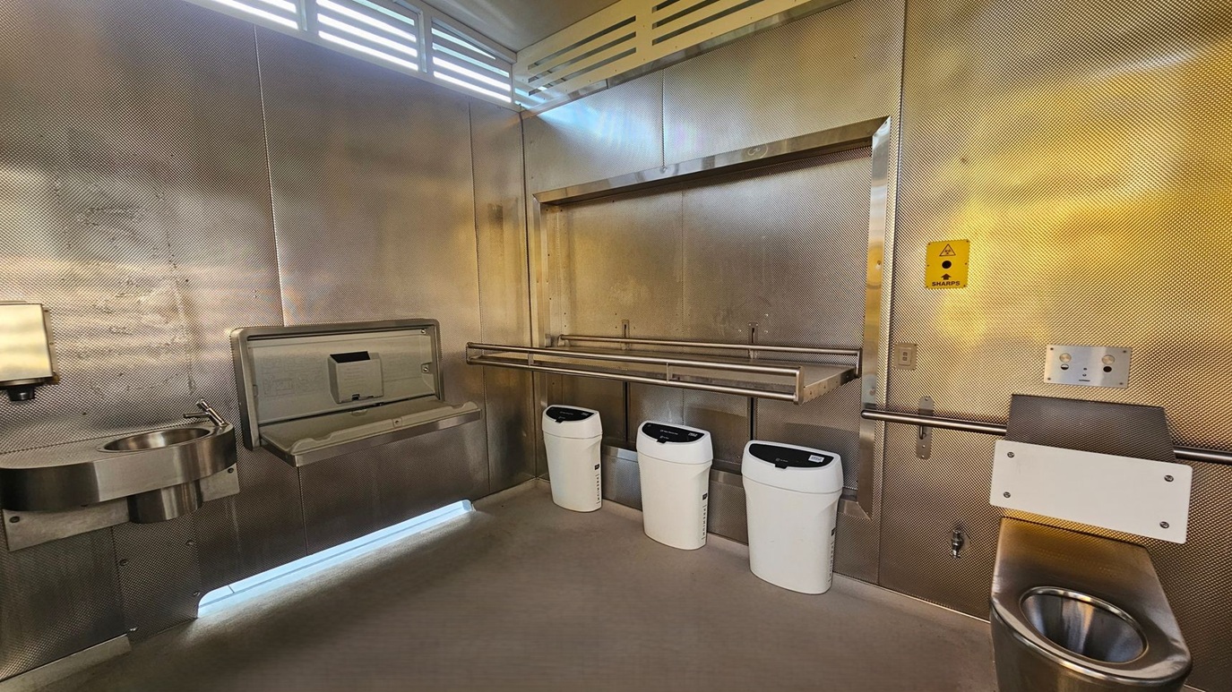 A public restroom with metallic walls and a concrete floor. On the left, there's a stainless steel sink with a soap dispenser above it. Next to the sink is a baby changing station. In the centre, three white bins are mounted on the wall. On the right, a stainless steel toilet with a safety rail and a small control panel above it. A yellow sharps disposal sign is visible on the wall. The ceiling has a vented section allowing light to filter through. A public restroom with metallic walls and a concrete floor. On the left, there's a stainless steel sink with a soap dispenser above it. Next to the sink is a baby changing station. In the centre, three white bins are mounted on the wall. On the right, a stainless steel toilet with a safety rail and a small control panel above it. A yellow sharps disposal sign is visible on the wall. The ceiling has a vented section allowing light to filter through.