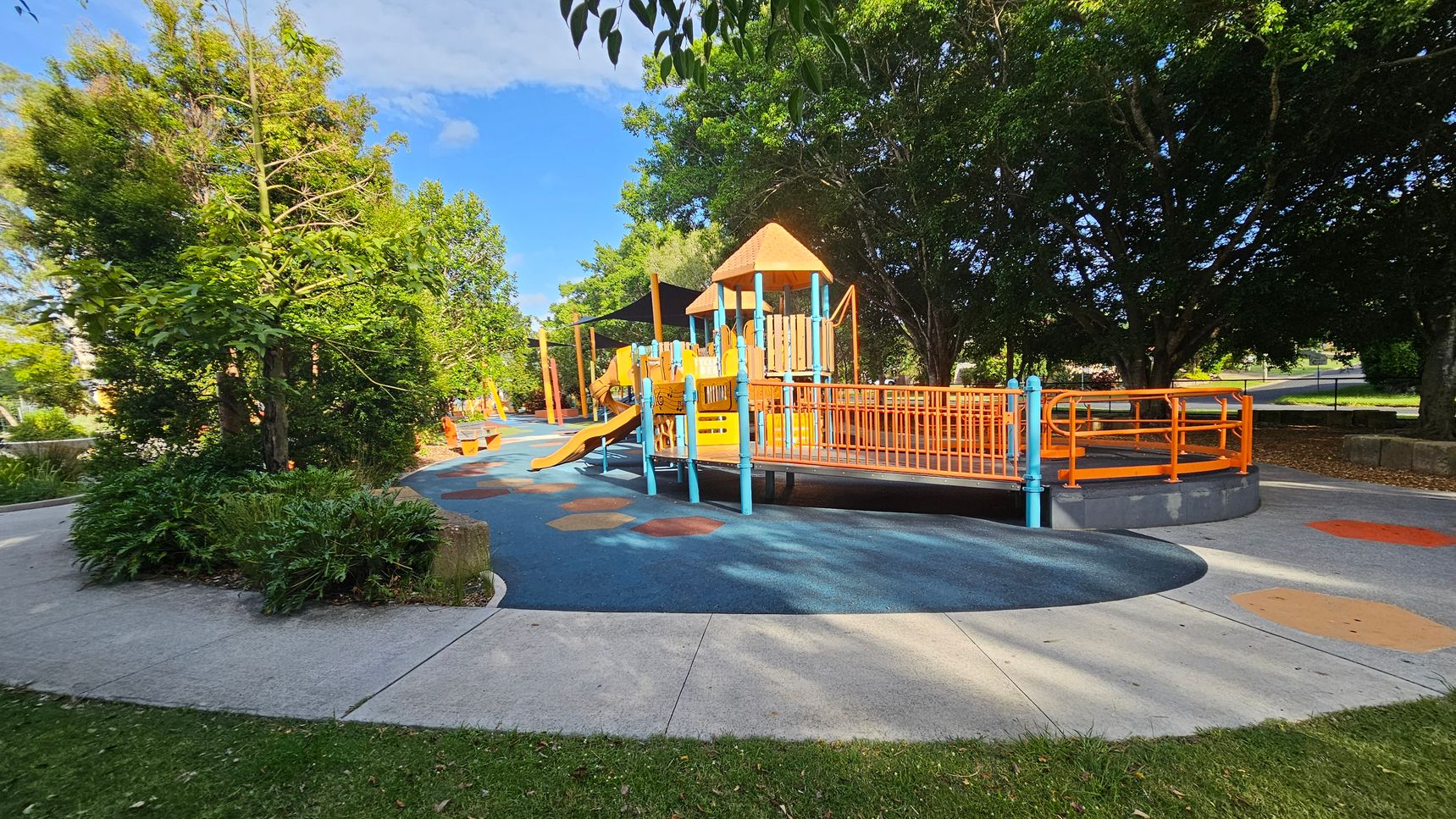 A playground with a colourful play structure featuring orange and blue elements, including slides and ramps, is in the centre. The ground is covered with a blue and orange rubber surface. On the left, there are lush green trees and shrubs. In the background, more trees provide shade. The foreground has a concrete path and grass. A playground with a colourful play structure featuring orange and blue elements, including slides and ramps, is in the centre. The ground is covered with a blue and orange rubber surface. On the left, there are lush green trees and shrubs. In the background, more trees provide shade. The foreground has a concrete path and grass.