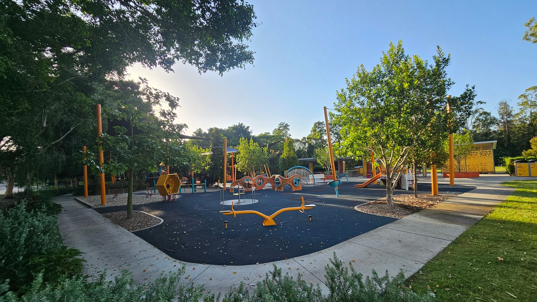 A playground with various equipment is surrounded by trees and greenery. On the left, there are large trees providing shade. In the centre, a black rubber play surface features a yellow seesaw, a hexagonal climbing structure, and other play equipment. To the right, more trees and a slide are visible. The ground is a mix of concrete paths and grass, with some mulch around the trees. The background includes more trees and a yellow structure. A playground with various equipment is surrounded by trees and greenery. On the left, there are large trees providing shade. In the centre, a black rubber play surface features a yellow seesaw, a hexagonal climbing structure, and other play equipment. To the right, more trees and a slide are visible. The ground is a mix of concrete paths and grass, with some mulch around the trees. The background includes more trees and a yellow structure.