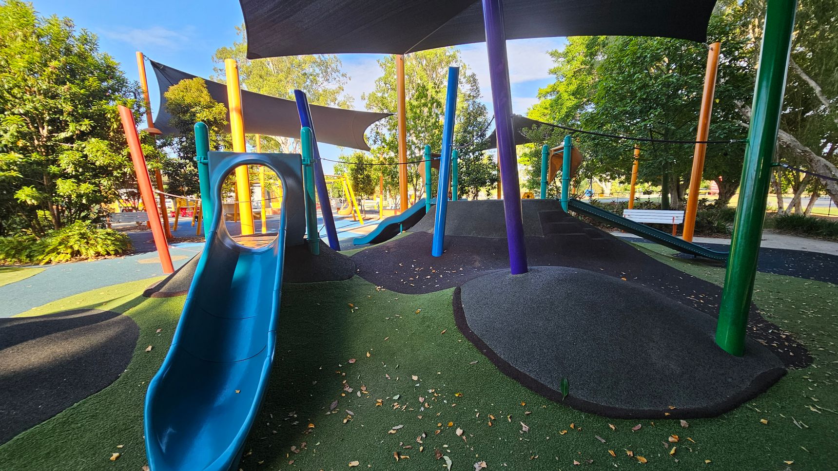 A playground with a blue slide on the left, leading down to a surface covered in green and black rubber. Colourful poles in blue, orange, and green support overhead shade sails. In the background, there are trees and additional playground equipment, including swings. A white bench is visible to the right, surrounded by more trees and foliage. The ground is scattered with fallen leaves. A playground with a blue slide on the left, leading down to a surface covered in green and black rubber. Colourful poles in blue, orange, and green support overhead shade sails. In the background, there are trees and additional playground equipment, including swings. A white bench is visible to the right, surrounded by more trees and foliage. The ground is scattered with fallen leaves.