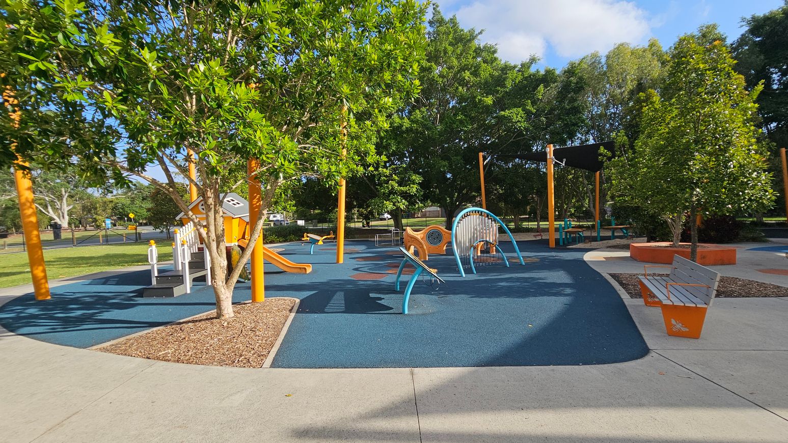A playground with various equipment on a blue rubber surface. On the left, there's a tree with green leaves and an orange slide. In the centre, a climbing structure with blue arches and a small orange playhouse. On the right, a bench with orange accents and another tree. The background features more trees and a shaded area with a black canopy. The ground is a mix of concrete paths and mulch around the trees. A playground with various equipment on a blue rubber surface. On the left, there's a tree with green leaves and an orange slide. In the centre, a climbing structure with blue arches and a small orange playhouse. On the right, a bench with orange accents and another tree. The background features more trees and a shaded area with a black canopy. The ground is a mix of concrete paths and mulch around the trees.