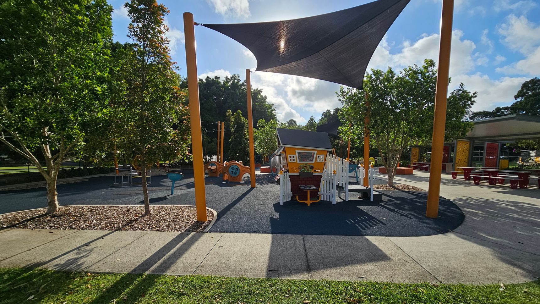 A playground with a small playhouse in the centre, surrounded by a white picket fence. The ground is covered with a soft, dark surface. Tall orange poles support a large triangular shade sail overhead. To the left, there are trees and a play structure with a blue seat. On the right, picnic tables are visible under a shelter. The foreground has a grassy area and a concrete path, while more trees and playground equipment are in the background. A playground with a small playhouse in the centre, surrounded by a white picket fence. The ground is covered with a soft, dark surface. Tall orange poles support a large triangular shade sail overhead. To the left, there are trees and a play structure with a blue seat. On the right, picnic tables are visible under a shelter. The foreground has a grassy area and a concrete path, while more trees and playground equipment are in the background.