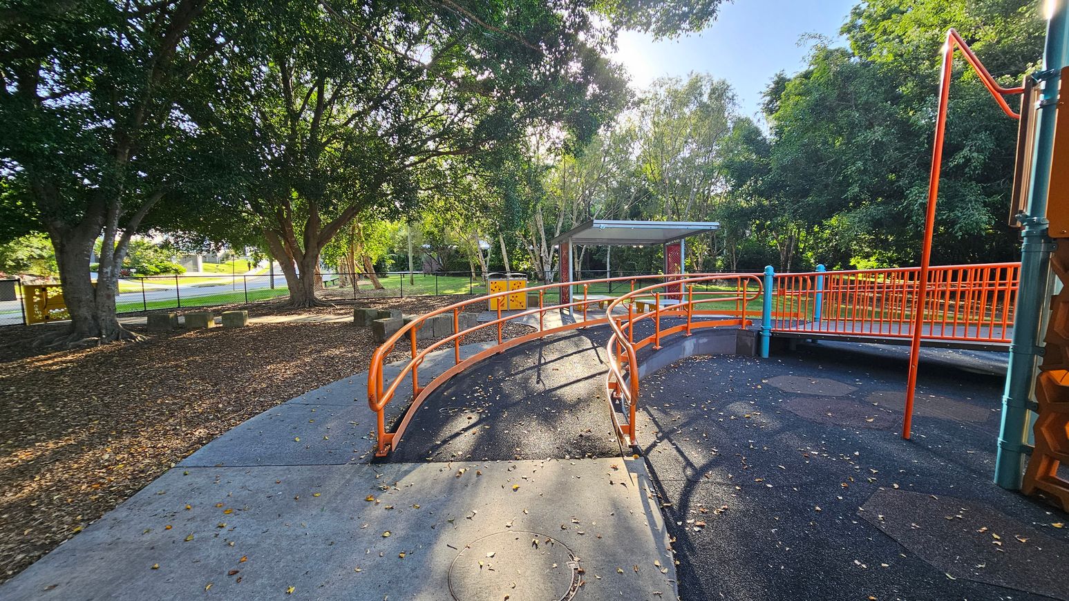 A playground with an orange railing bridge in the centre, leading to a covered area in the background. The ground is a mix of concrete and rubberised surface with scattered leaves. On the left, large trees provide shade, and a fence runs along the perimeter. On the right, more playground equipment is partially visible. The area is surrounded by greenery, creating a natural setting. A playground with an orange railing bridge in the centre, leading to a covered area in the background. The ground is a mix of concrete and rubberised surface with scattered leaves. On the left, large trees provide shade, and a fence runs along the perimeter. On the right, more playground equipment is partially visible. The area is surrounded by greenery, creating a natural setting.
