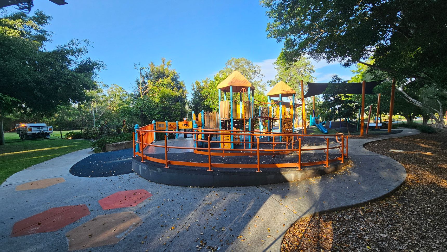 A playground with colourful equipment featuring orange and blue structures, including slides and climbing frames, is in the centre. The ground is a mix of concrete and rubber surfaces with geometric patterns. Trees surround the area, providing shade, and a path curves around the playground. A white utility vehicle is parked on the grass to the left. The background is filled with lush greenery and more trees. A playground with colourful equipment featuring orange and blue structures, including slides and climbing frames, is in the centre. The ground is a mix of concrete and rubber surfaces with geometric patterns. Trees surround the area, providing shade, and a path curves around the playground. A white utility vehicle is parked on the grass to the left. The background is filled with lush greenery and more trees.