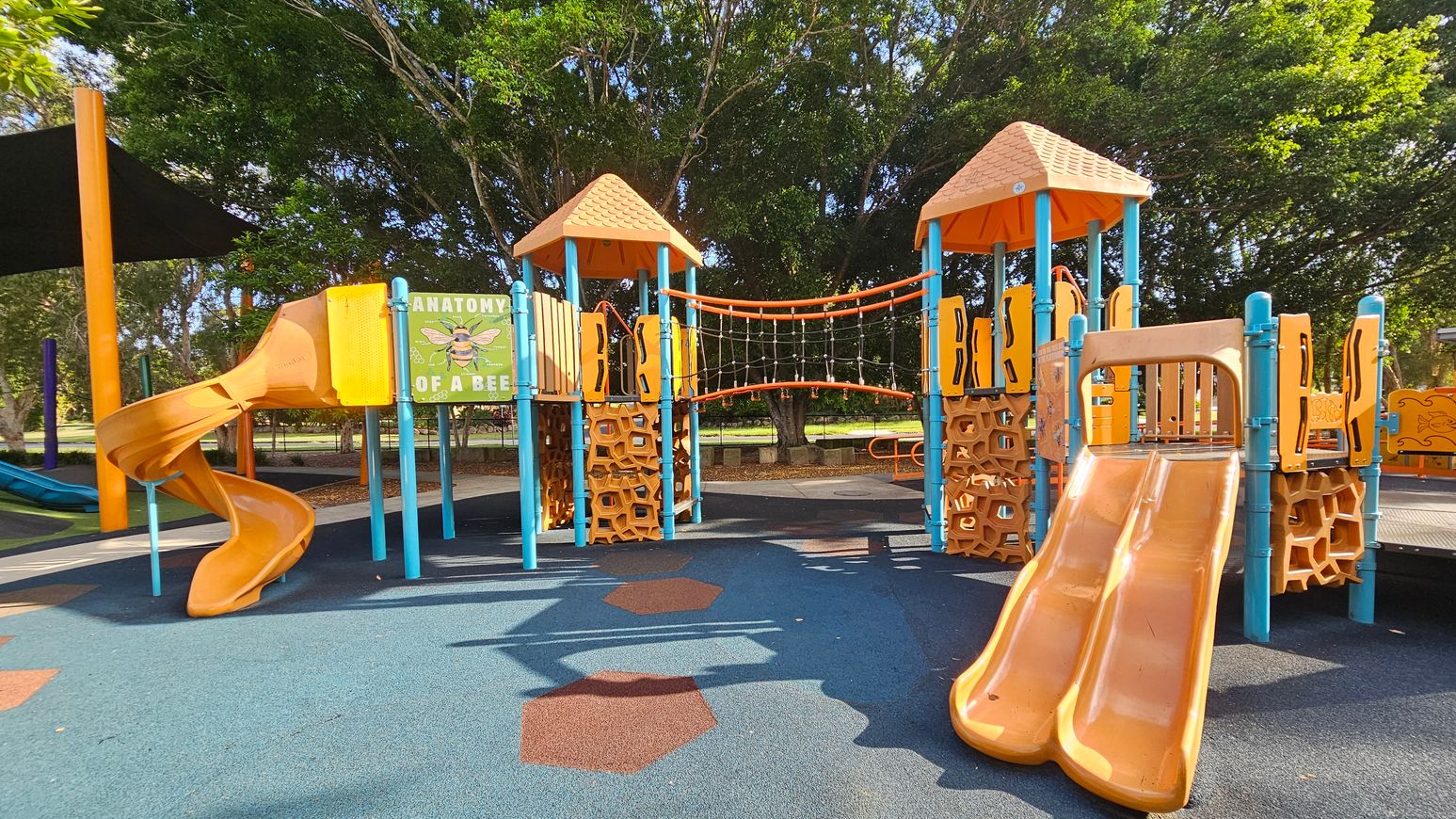 A children's playground with two orange slides, one spiral on the left and a double slide on the right. The structures are supported by blue poles and feature orange hexagonal patterns. A sign titled "Anatomy of a Bee" is visible on the left. An orange rope bridge connects two towers with orange roofs. The ground is covered with blue and orange rubber tiles. Trees are in the background, providing shade. A children's playground with two orange slides, one spiral on the left and a double slide on the right. The structures are supported by blue poles and feature orange hexagonal patterns. A sign titled "Anatomy of a Bee" is visible on the left. An orange rope bridge connects two towers with orange roofs. The ground is covered with blue and orange rubber tiles. Trees are in the background, providing shade.