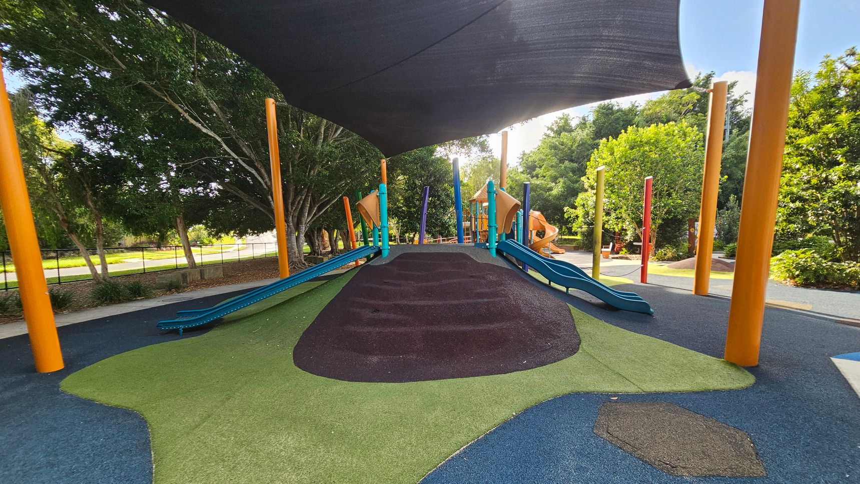 A playground with a central mound covered in green and brown rubber surfacing, flanked by blue slides. Bright orange and blue poles support a large black shade canopy overhead. In the background, there are trees and additional playground equipment, including orange slides. The ground is a mix of blue and green rubberised material, and the area is surrounded by lush greenery. A playground with a central mound covered in green and brown rubber surfacing, flanked by blue slides. Bright orange and blue poles support a large black shade canopy overhead. In the background, there are trees and additional playground equipment, including orange slides. The ground is a mix of blue and green rubberised material, and the area is surrounded by lush greenery.