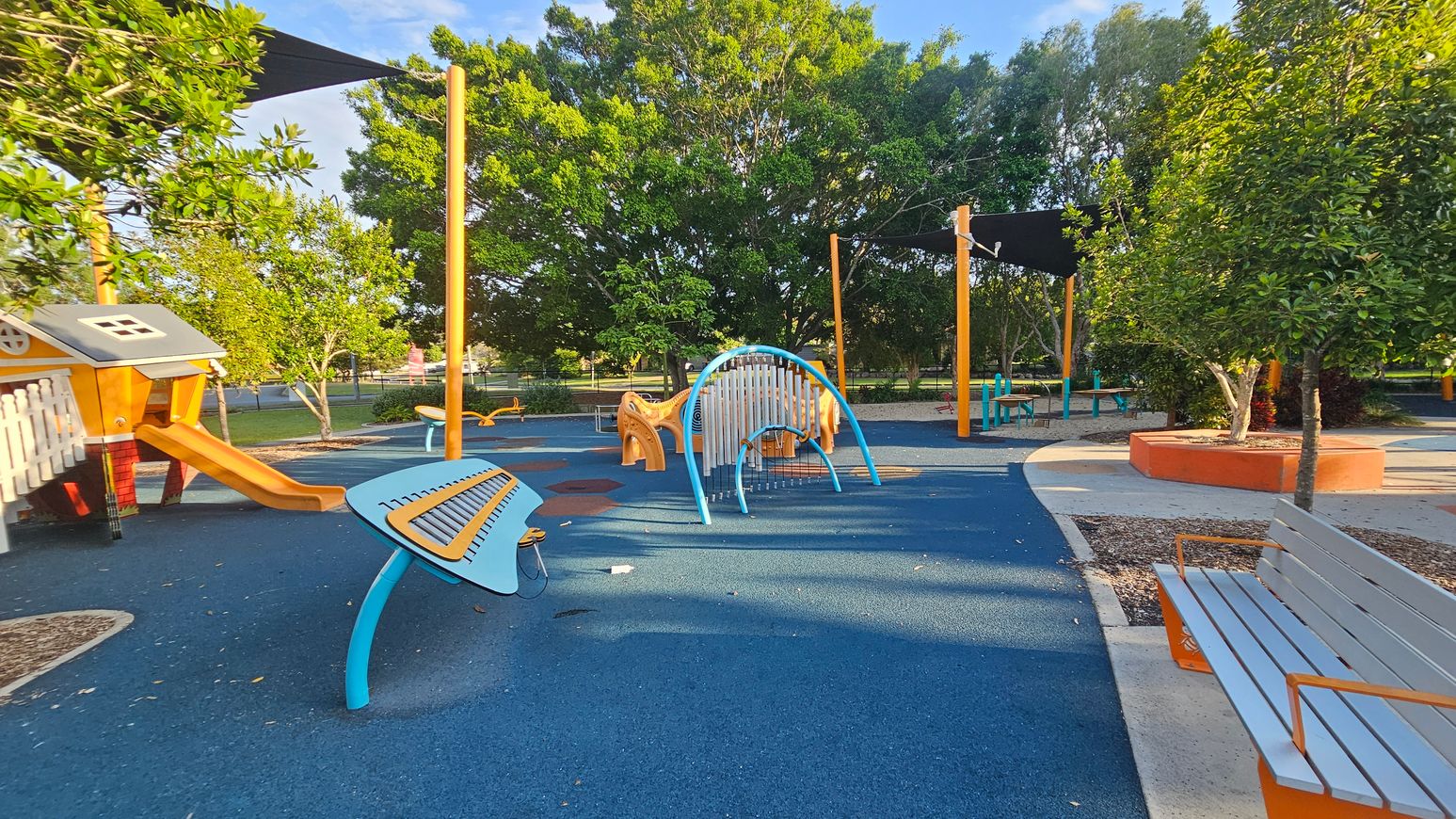 A playground with a blue rubber floor. On the left, there's a playhouse with a slide and a musical play structure. In the centre, a curved climbing frame is visible. To the right, a bench with orange supports is near a tree planted in a circular orange planter. In the background, there are more trees and a shaded area with additional seating. A playground with a blue rubber floor. On the left, there's a playhouse with a slide and a musical play structure. In the centre, a curved climbing frame is visible. To the right, a bench with orange supports is near a tree planted in a circular orange planter. In the background, there are more trees and a shaded area with additional seating.
