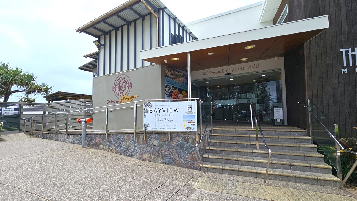 Stepped entrance - Arrival and Parking - The Surf Club Mooloolaba. Entrance to a modern building with a sloped path and steps leading up to glass doors. On the left, a wall features a logo and a sign for "Bayview Bar & Bites." The building has a mix of stone and wood panelling, with a flat roof extending over the entrance. A railing lines the path and steps. To the left, there are trees and a wooden fence and ramp to the entrance