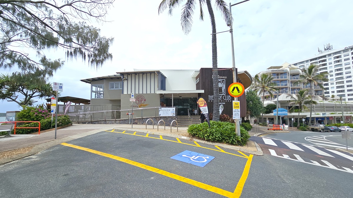 Accessible parking.  Arrival and parking. The Surf Club Mooloolaba. A modern building with a grey facade is in the centre, featuring a ramp and bike racks in front. To the right, a pedestrian crossing and a palm tree are visible. A sign with a pedestrian symbol is near the crossing. On the left, there are trees and a glimpse of the ocean. The foreground shows a marked parking space with yellow lines and a blue disabled symbol. In the background, a multi-storey building with balconies is visible.