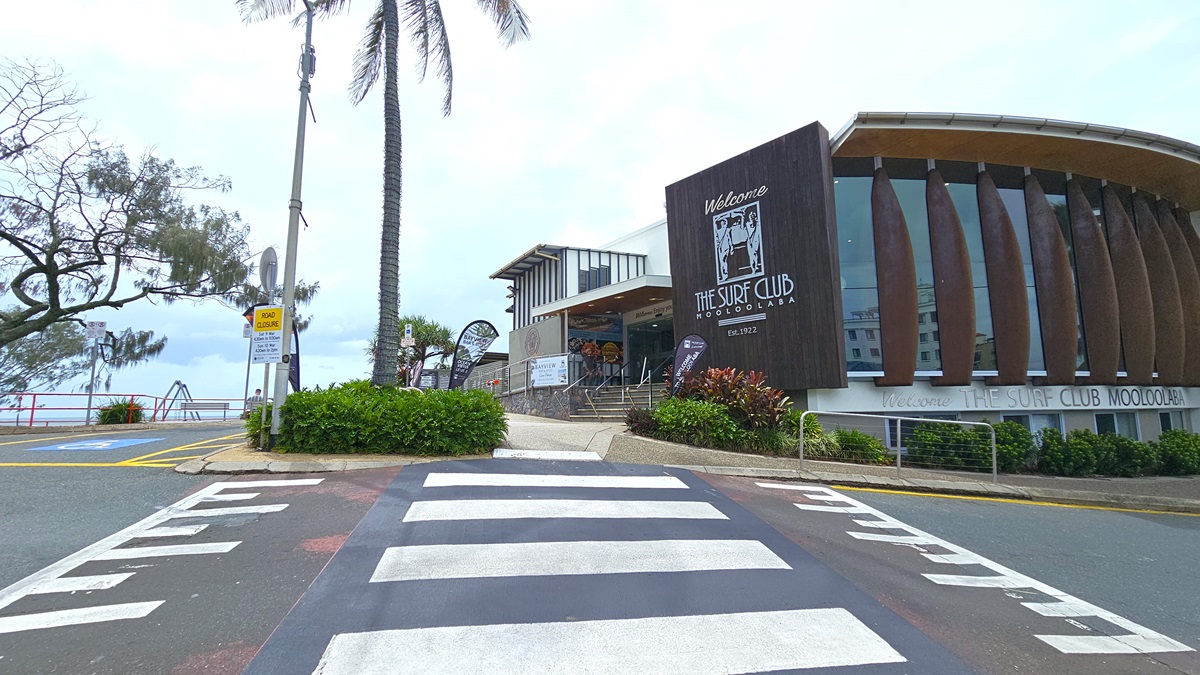 Pedestrian crossing - Arrival and parking - The Surf Club Mooloolaba. The building structure features wooden slats and large windows. A zebra crossing leads towards the entrance, which is flanked by greenery and a palm tree. On the left, there are more trees and a glimpse of the ocean with a railing. The pavement is marked with a pedestrian crossing, and there are signs and banners near the entrance.