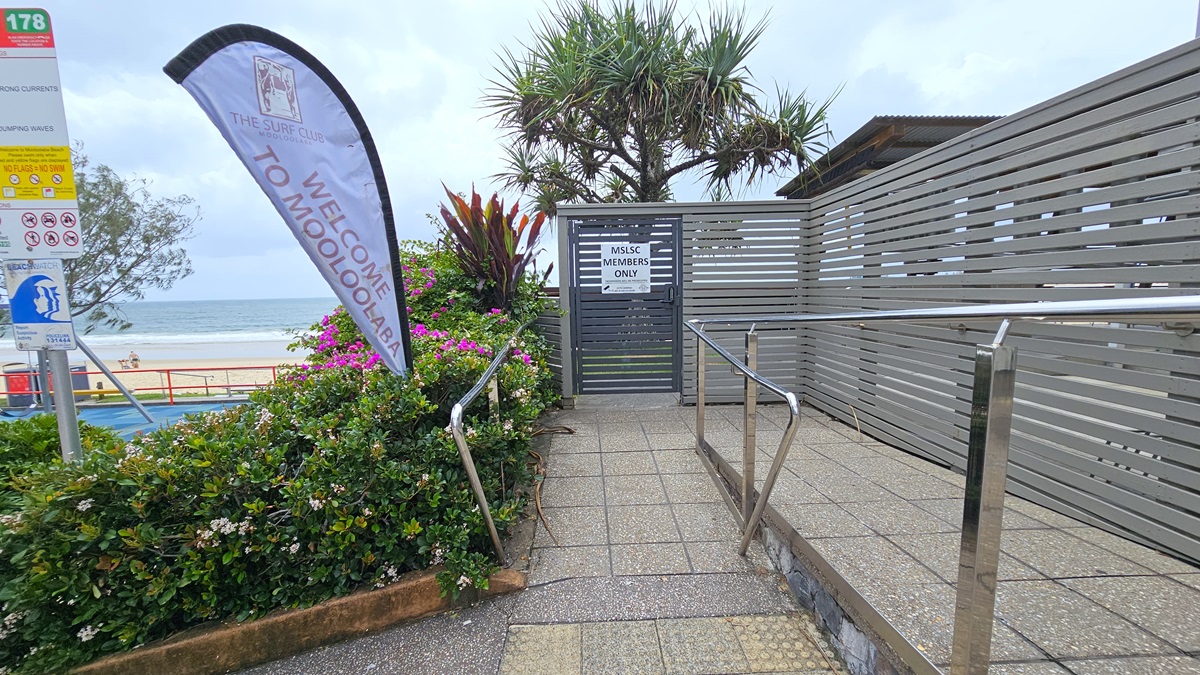 Accessible ramp entry - Arrival and parking - The Surf Club Mooloolaba. A pathway with metal railings leads to a gated entrance marked "MSLSC Members Only". On the left, a flag reads "Welcome to Mooloolaba" next to a signpost with various symbols. Lush greenery and pink flowers line the path. In the background, a beach and ocean are visible, with a tree providing some shade. The area is bordered by a wooden slat fence.