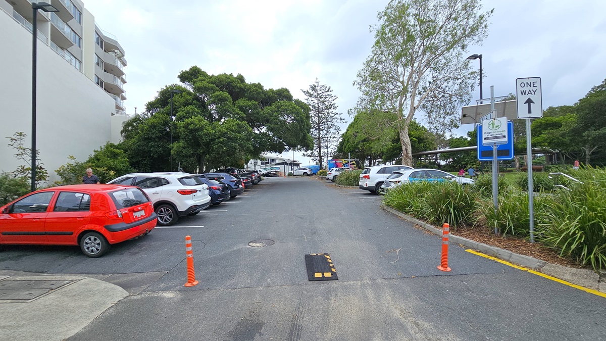General parking area - Arrival and Parking - The Surf Club Mooloolaba. A car park with vehicles lined up on both sides. On the left, a red car is parked near a tall building with balconies. On the right, a "One Way" sign is visible next to some greenery and a tree. The ground is paved, and there are orange traffic cones and a speed bump in the centre. In the background, more trees and parked cars are visible, with a glimpse of a structure further back.