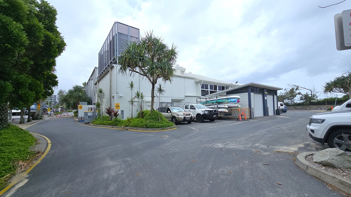 Street view - Arrival and parking - The Surf Club Mooloolaba. A street scene with a large white building on the left, featuring a modern design with vertical slats on the upper section. Several vehicles, including utes, are parked beside the building. In the foreground, a curved road with a yellow line runs alongside a landscaped area with trees and shrubs. On the right, a smaller grey building is visible, and a white SUV is partially seen.