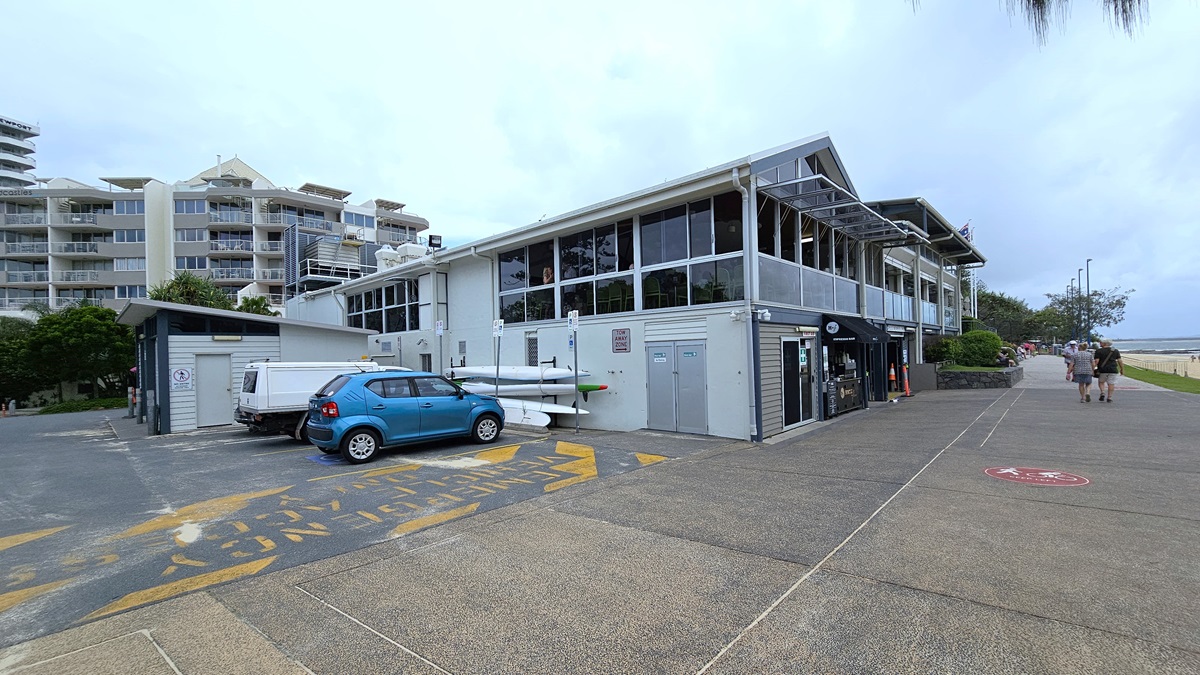 Building exterior and beach - Arrival and Parking - The Surf Club Mooloolaba. A two-storey building with large windows and a sloped roof is on the right. In front, a blue car and a white van are parked, with several kayaks stacked nearby. The pavement is marked with yellow and blue patterns. On the left, there are multi-storey residential buildings with balconies. A pathway runs alongside the building, with a few people walking towards the beach visible in the background.