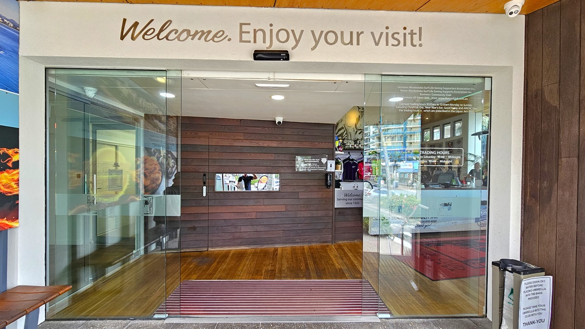Automatic sliding doors - Reception and Entrance - The Surf Club Mooloolaba. Glass entrance doors with "Welcome. Enjoy your visit!" written above. Inside, a wooden panelled wall is visible with a small window and some informational signs. To the right, a display with merchandise and brochures. The floor is wooden with a red mat near the entrance. A small bin is on the right outside the door. Ceiling lights are visible inside.