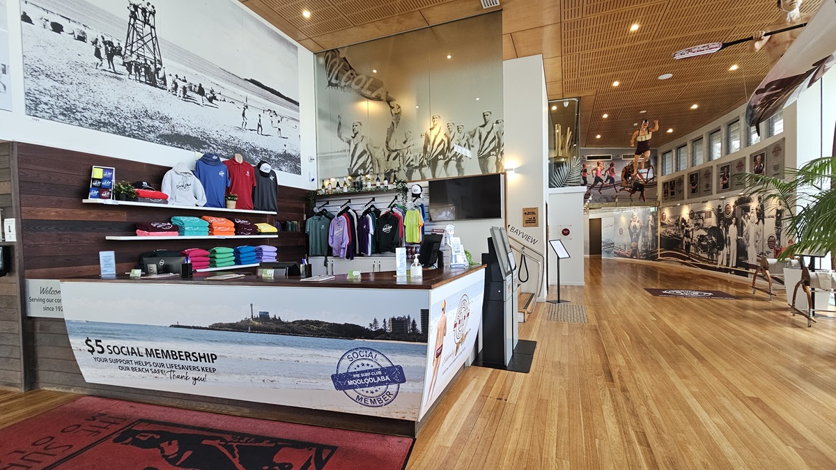 Reception Counter - Entrance and Reception - The Surf Club Mooloolaba. A spacious interior with a wooden floor and ceiling. On the left, a counter displays colourful folded shirts and hanging apparel. Behind the counter, a large black-and-white mural of a beach scene is visible. In the centre, a staircase leads to an upper level with more murals and mannequins dressed in sportswear. The right side features a long wall with historical photographs and a large plant.